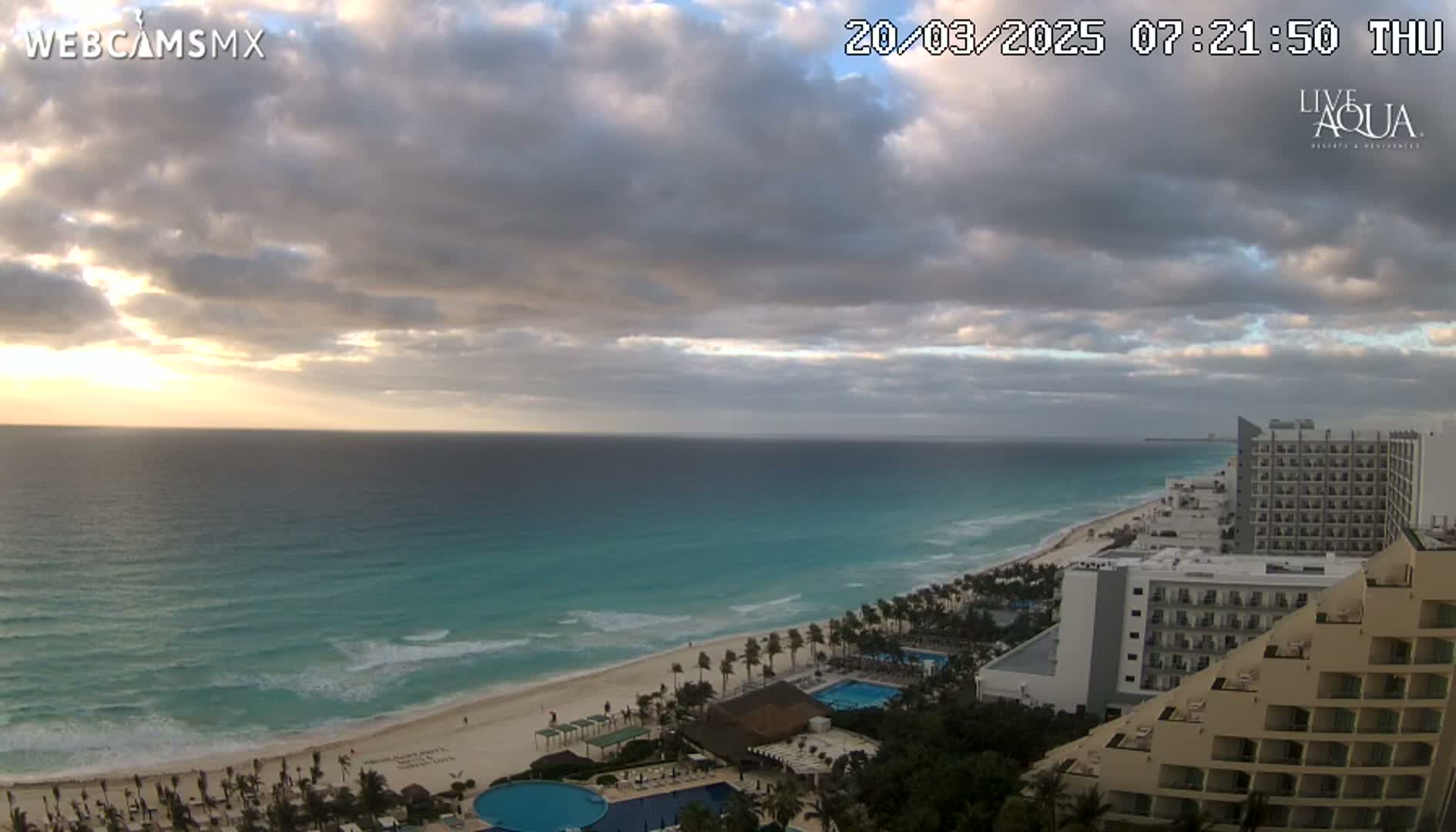 A partly cloudy sunrise over a turquoise ocean and sandy beach lined with resort hotels.
