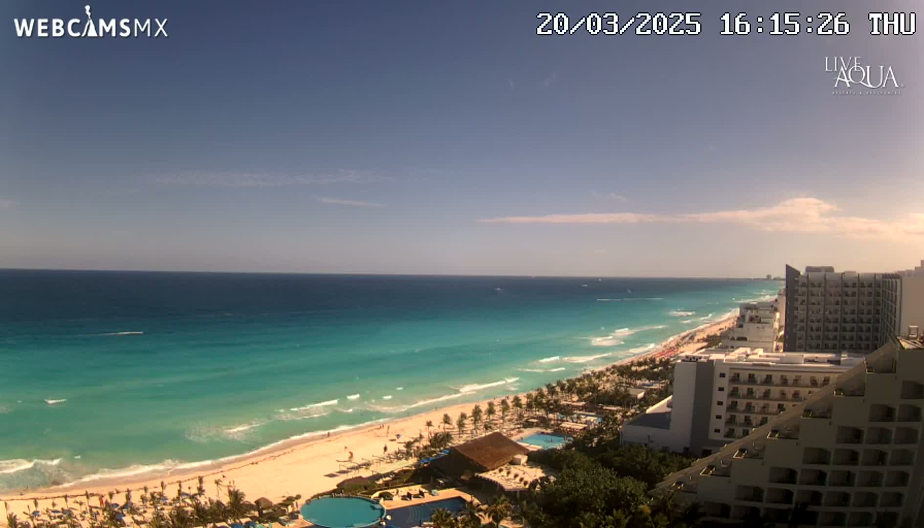 A mostly sunny day shows a turquoise ocean meeting a white-sand beach lined with hotels and palm trees.