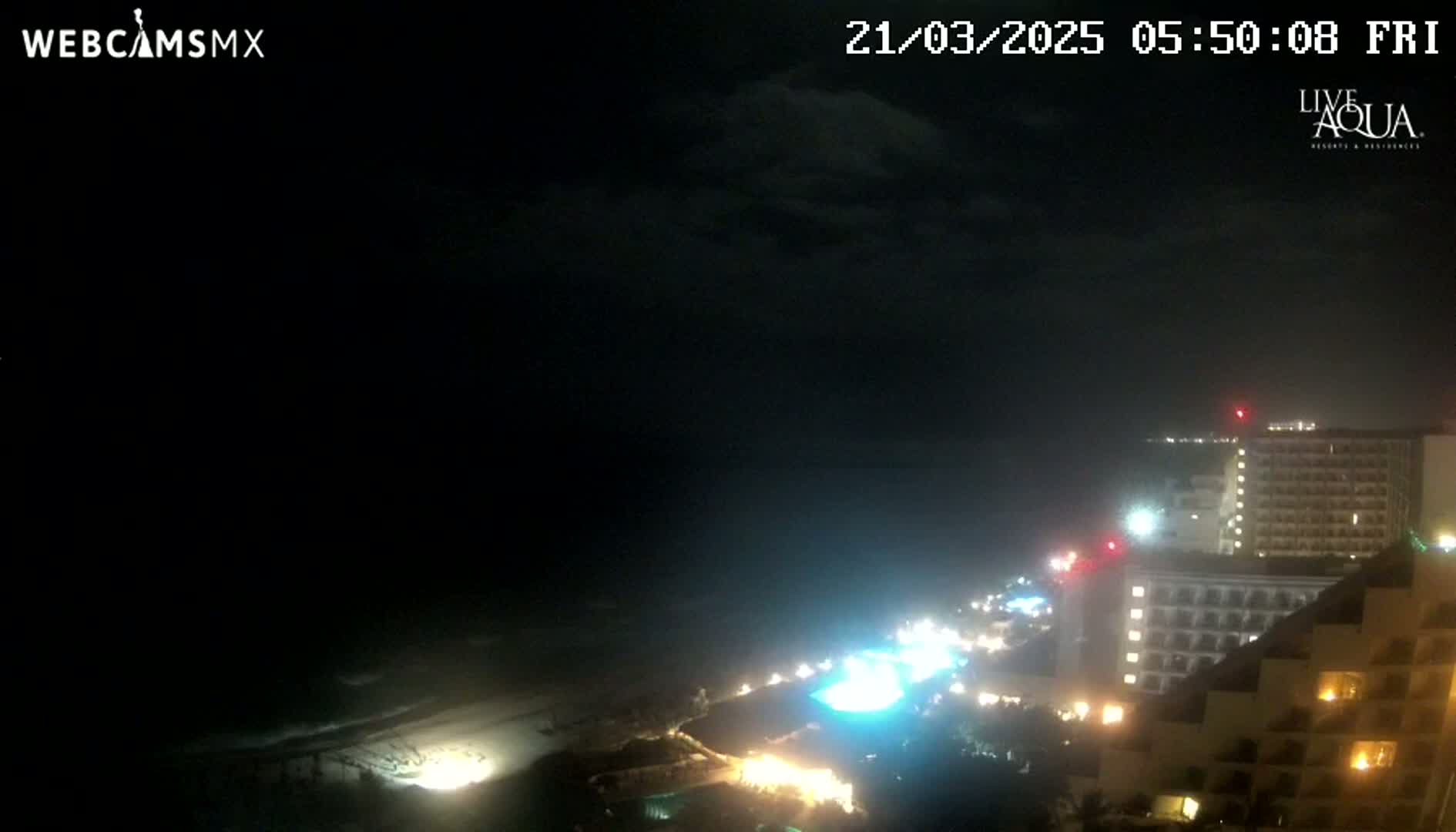 A nighttime view of a beach with illuminated buildings and a dark, cloudy sky.