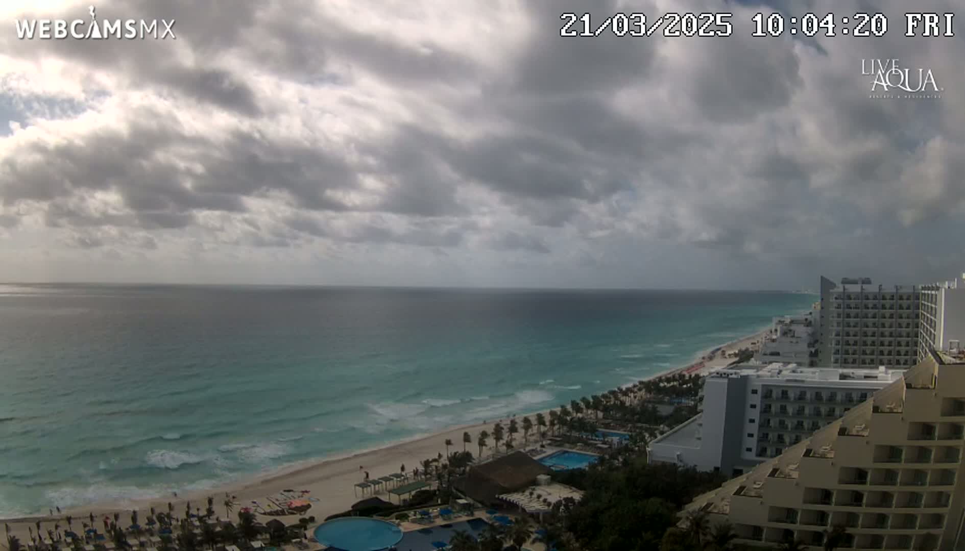 An ocean beach with turquoise water and cloudy skies is viewed from above, showing hotels and resorts lining the coast.