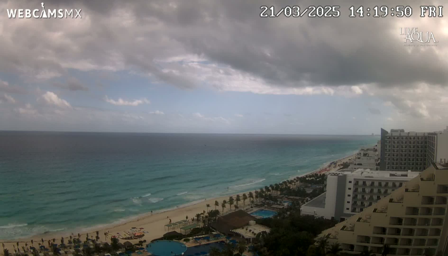 A partly cloudy sky overlooks a beach with turquoise water and several resort hotels.