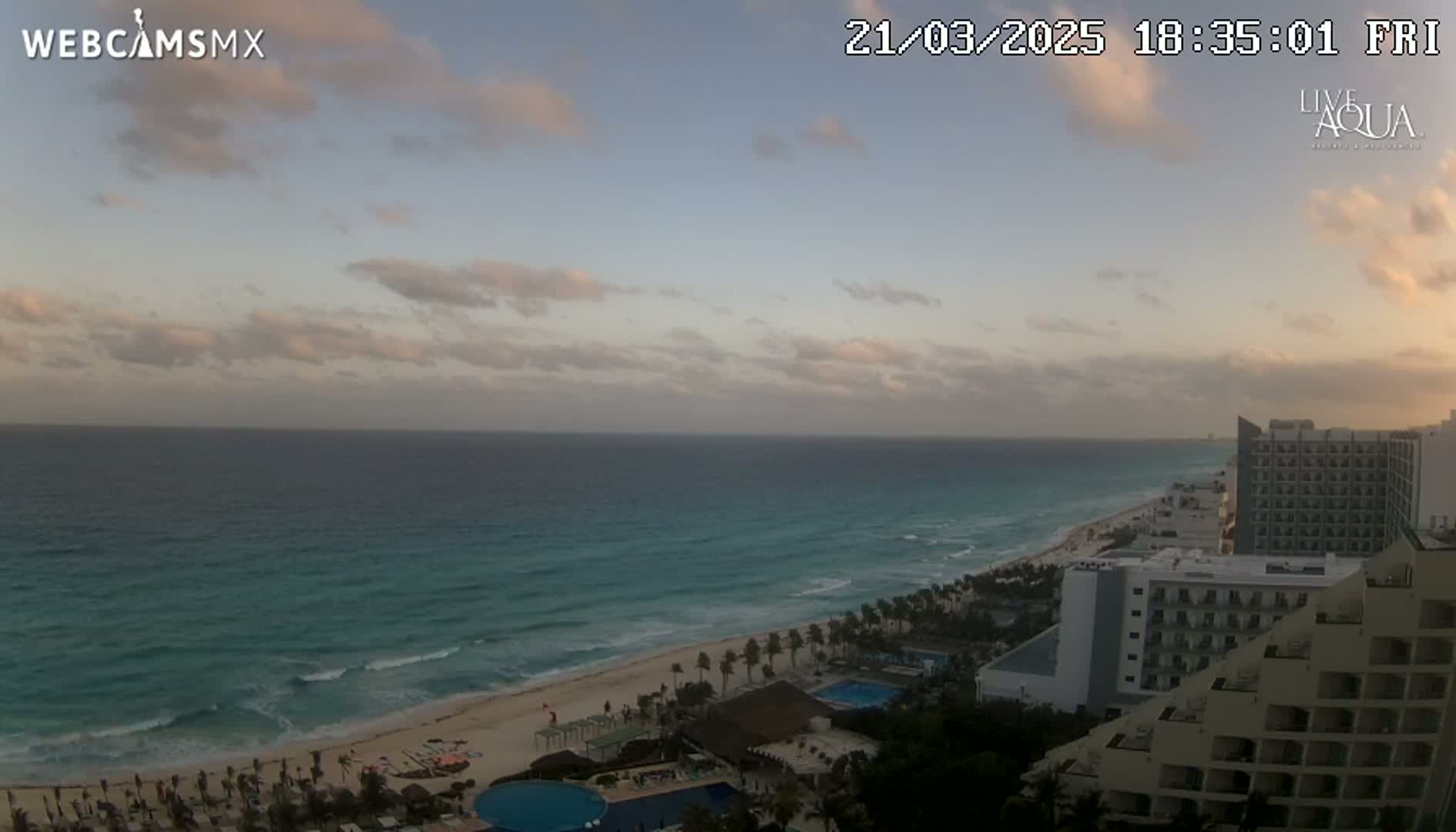 A mostly cloudy sunset over a beach with turquoise water and several hotels along the shore.
