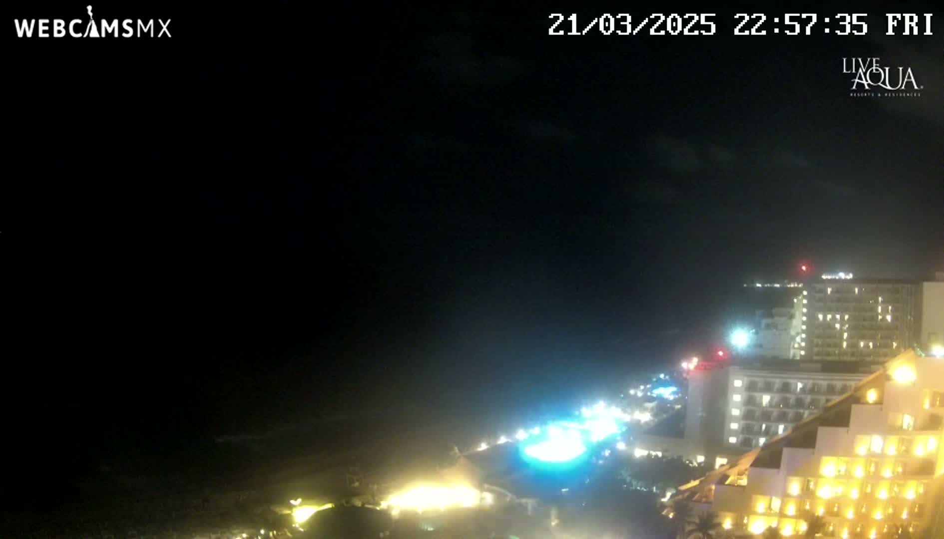 A nighttime coastal scene shows a dark ocean, a brightly lit beachfront area with several hotels, and a mostly clear sky.