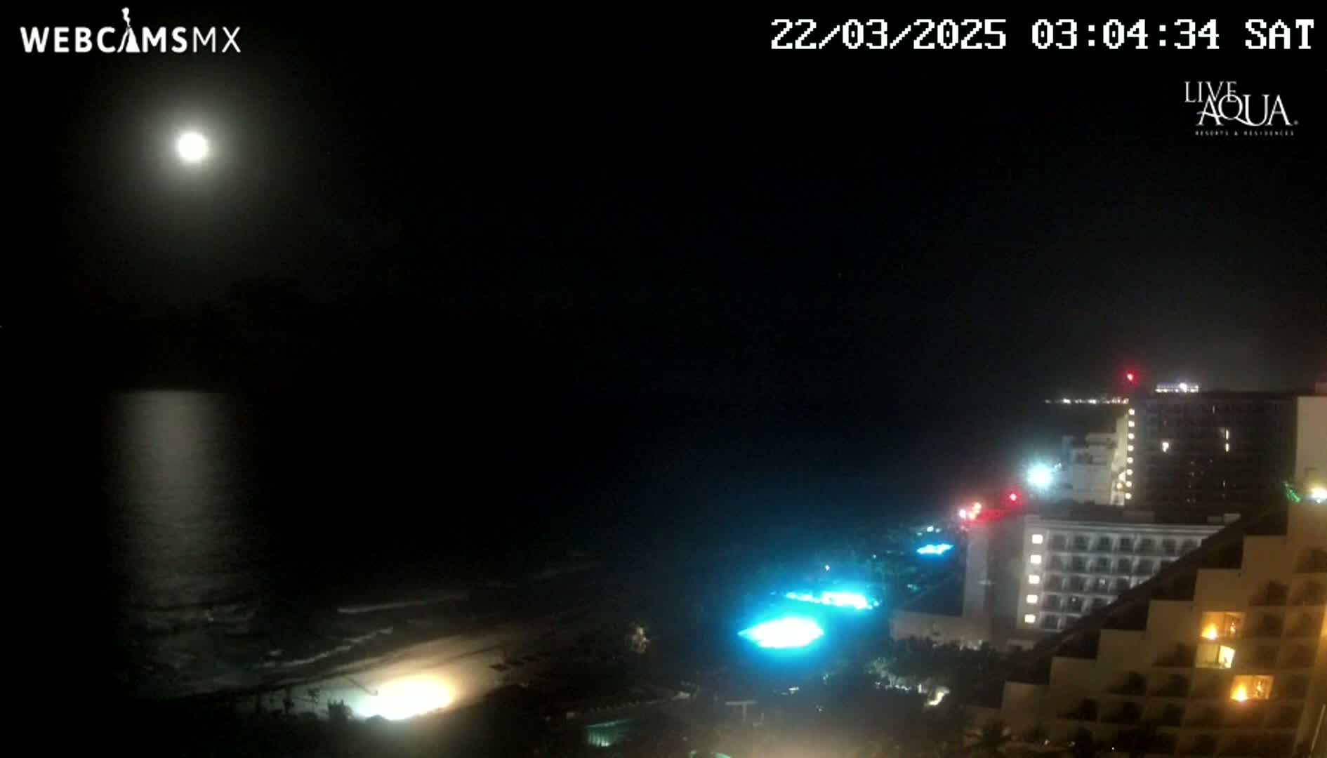 A bright moon illuminates a dark beach and ocean at night, with a large hotel complex visible in the foreground.