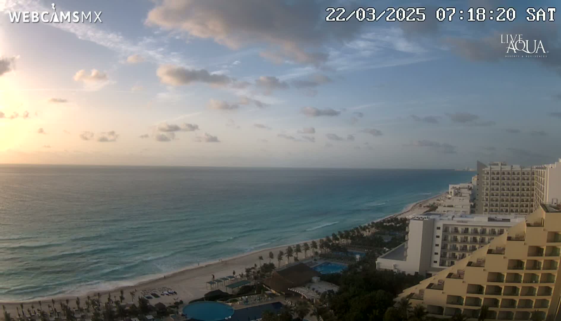 A partly cloudy sunrise casts a warm glow over a calm ocean, white sandy beach, and a line of beachfront hotels.