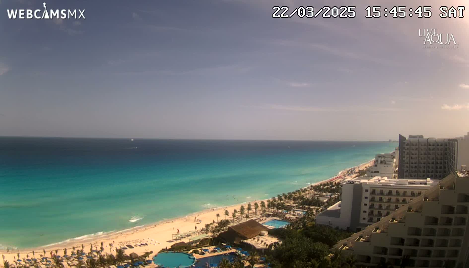 A mostly sunny day overlooking a long stretch of turquoise ocean meeting a white sandy beach lined with hotels and palm trees.
