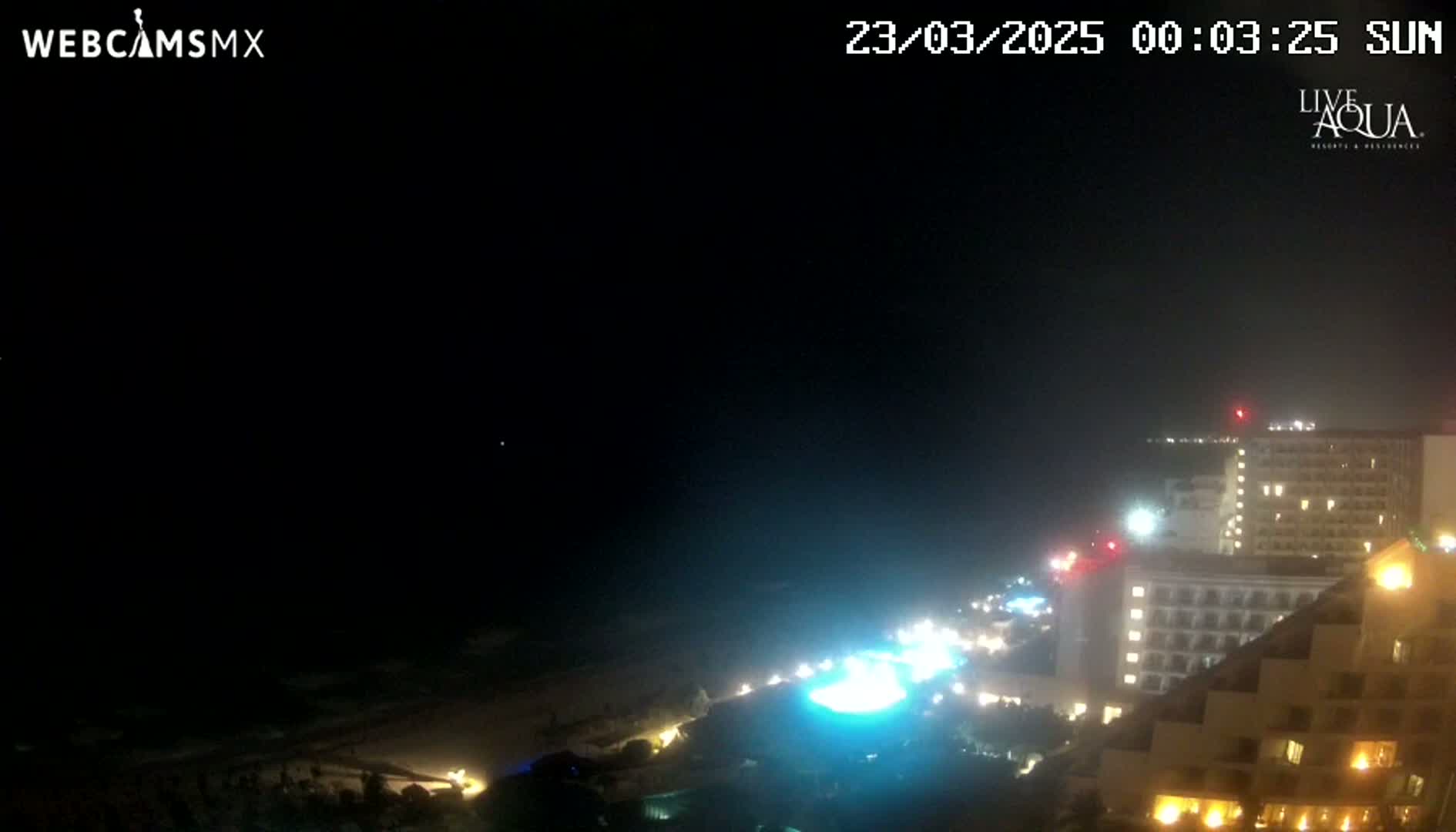 A nighttime, seaside view shows a brightly lit hotel complex next to a dark beach under a clear sky.