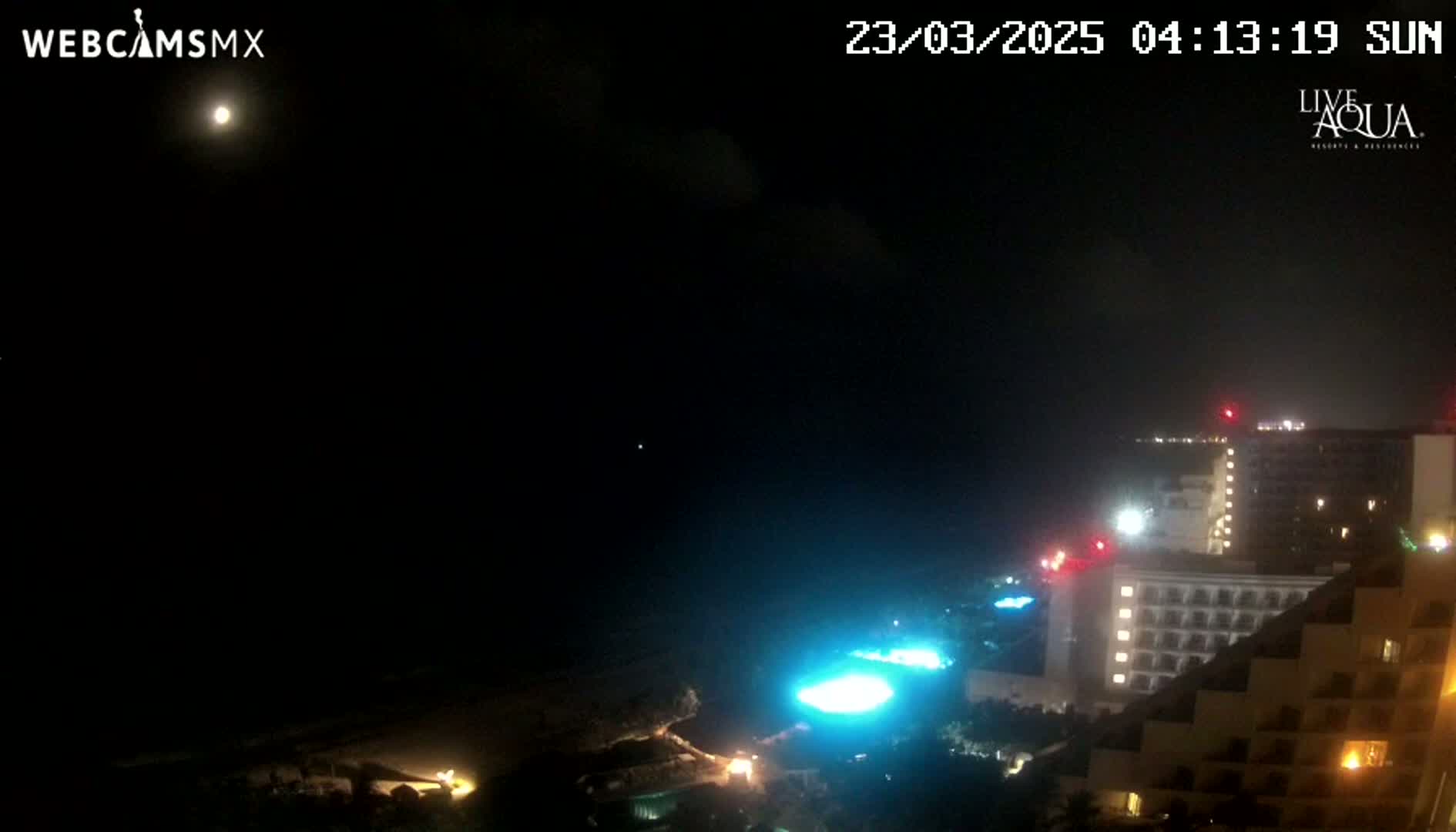 A nighttime view of a beachside resort illuminated under a partly cloudy sky with a bright moon visible.