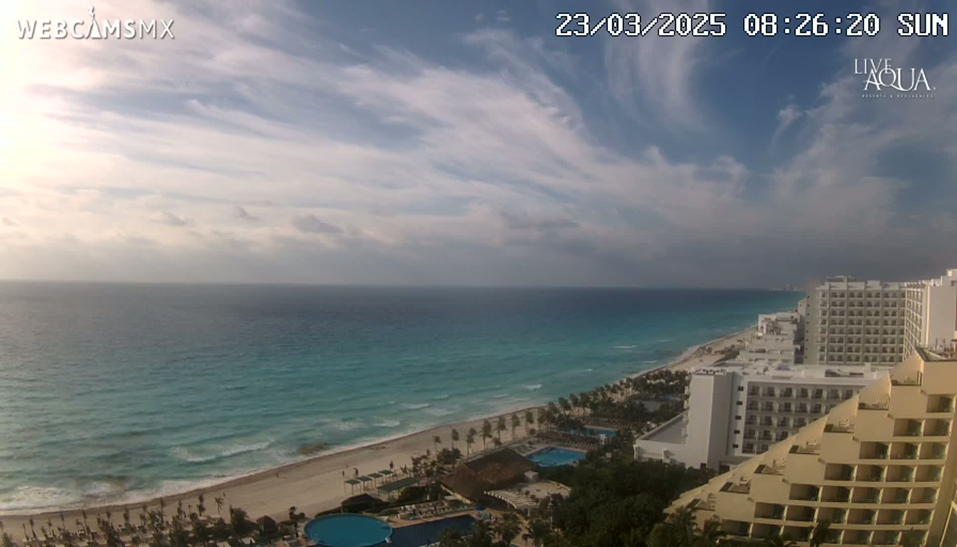 A partly cloudy sunny day shows a long stretch of beach and turquoise ocean next to a line of hotels.