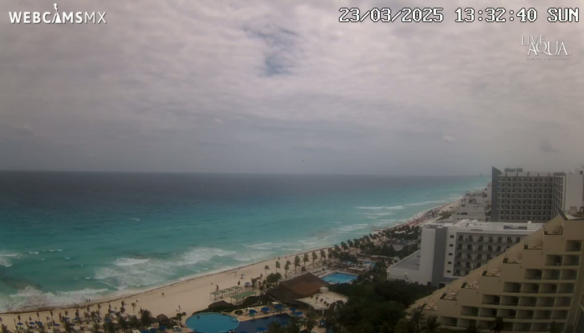 A mostly cloudy day reveals a beach with turquoise water and waves lapping a sandy shore, lined with resort hotels and swimming pools.