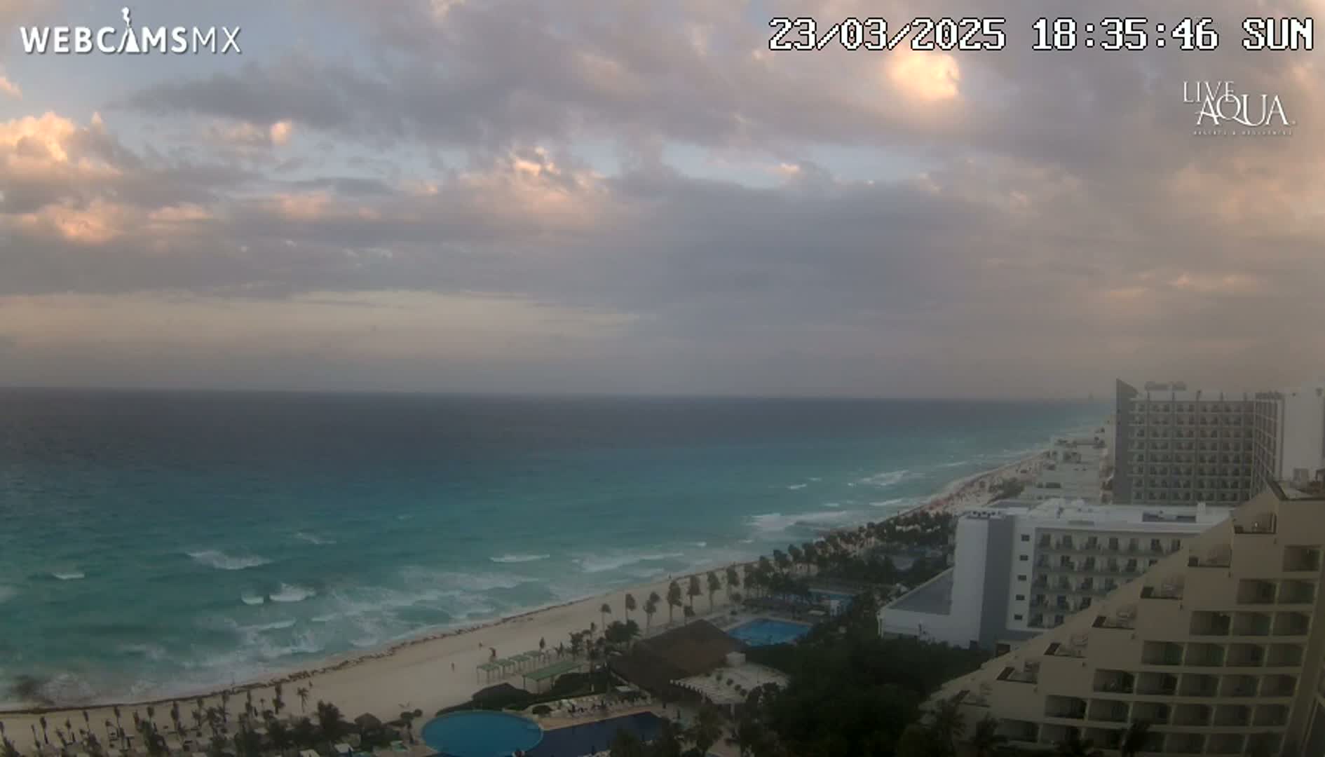A partly cloudy sunset over a turquoise ocean with waves breaking on a sandy beach, overlooking resort hotels.