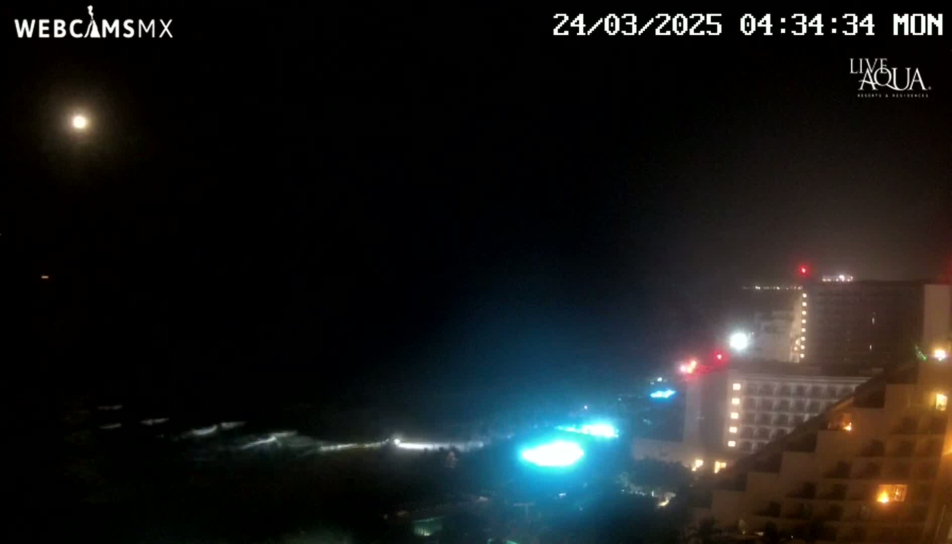 A nighttime coastal scene shows dark ocean waves gently lapping the shore under a mostly clear, moonlit sky, with illuminated buildings visible in the distance.