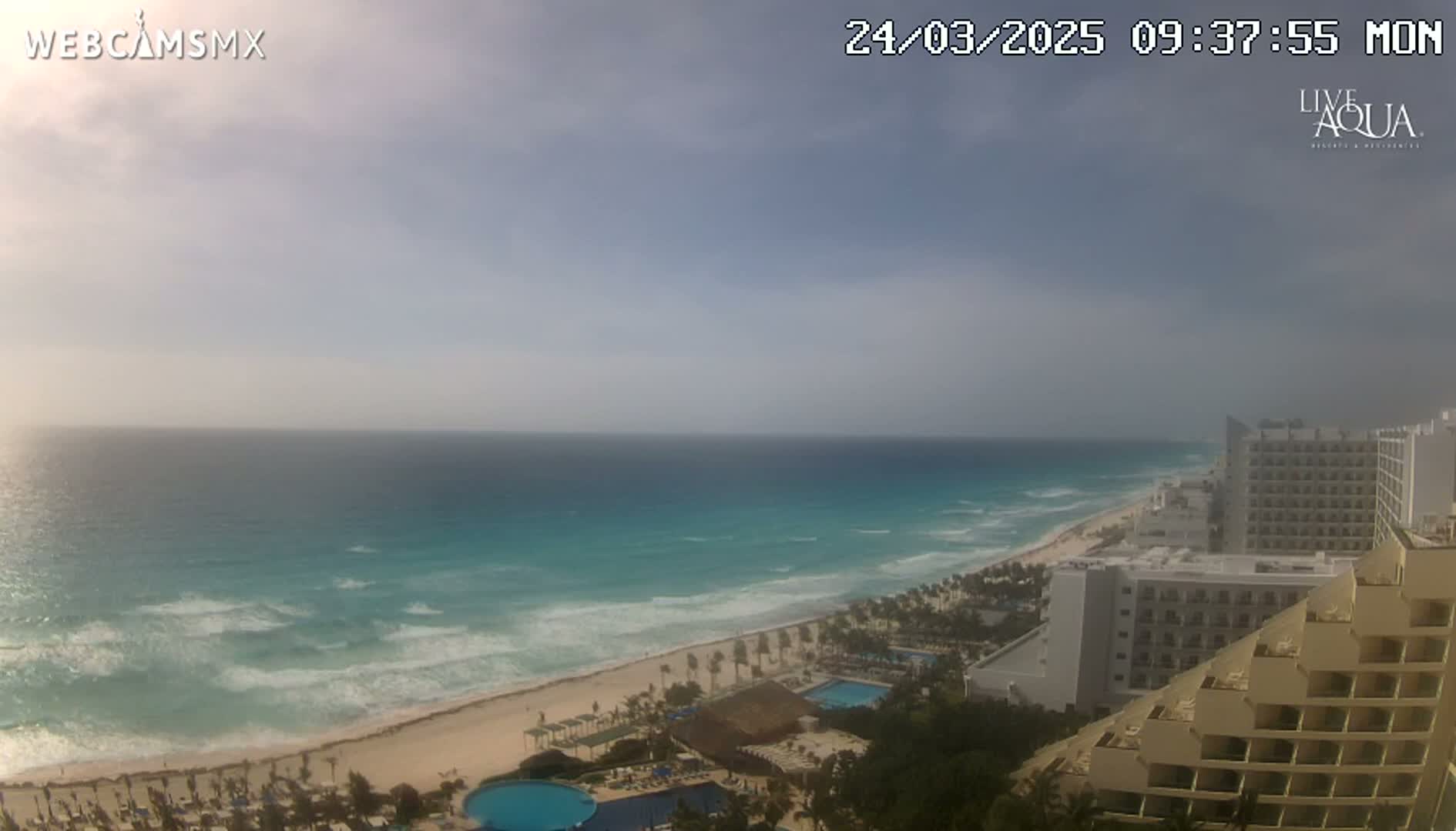 A mostly cloudy day shows a view of a beach with turquoise water, waves, and hotels along the coast.