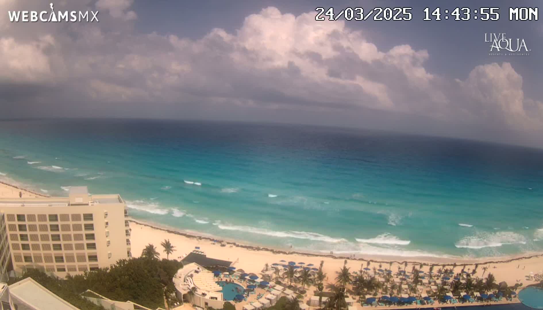 A partly cloudy day shows turquoise water lapping a sandy beach lined with umbrellas and a high-rise hotel.