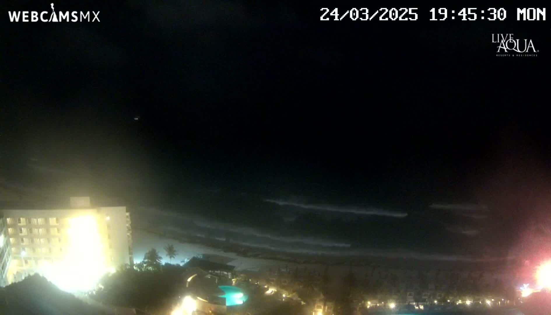 A nighttime view of a beach with gentle waves under a dark sky shows a brightly lit building to the left and a resort area below.