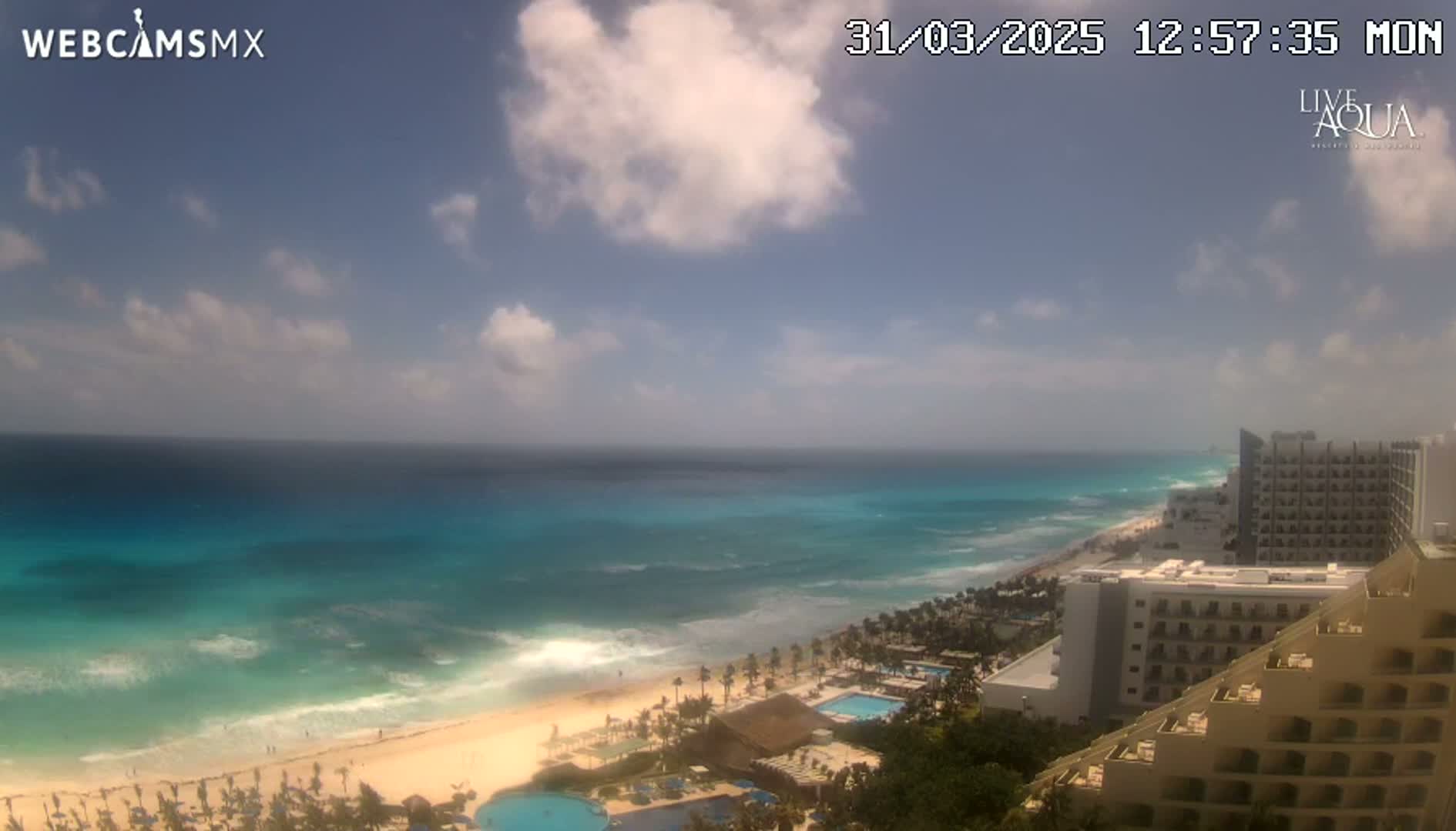A partly cloudy day shows a beach with turquoise water, waves, and a line of hotels along the coast.