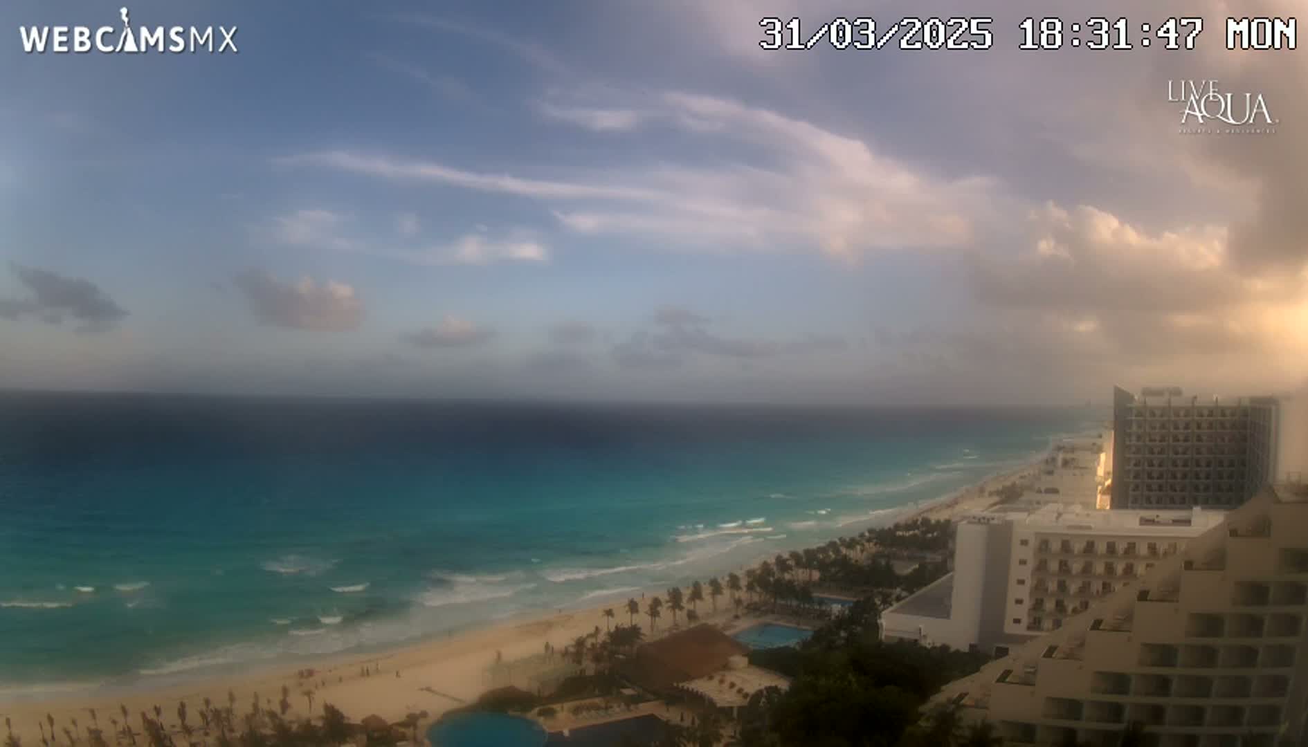 A partly cloudy sunset over a beach with turquoise water and beachfront hotels.
