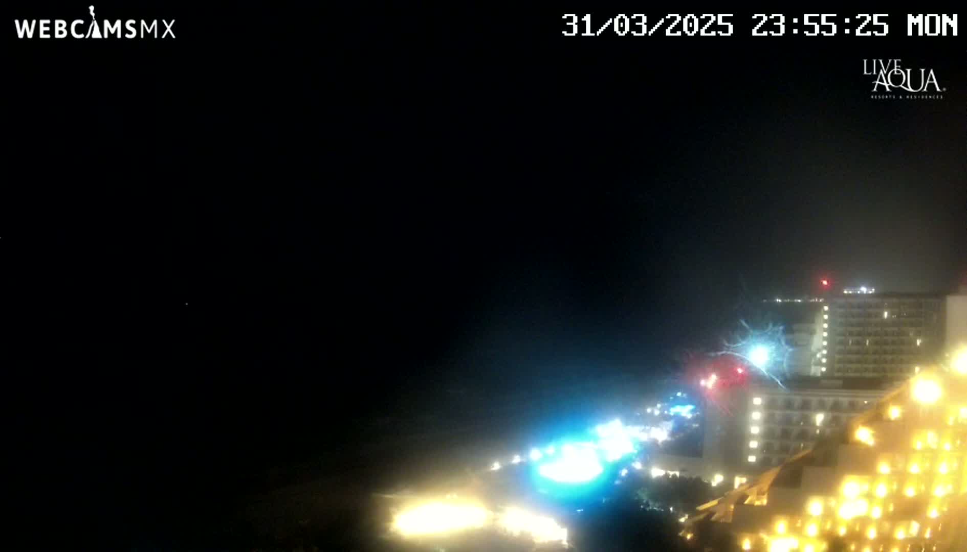 A nighttime view of brightly lit buildings near a coastline under a dark, possibly stormy, sky.