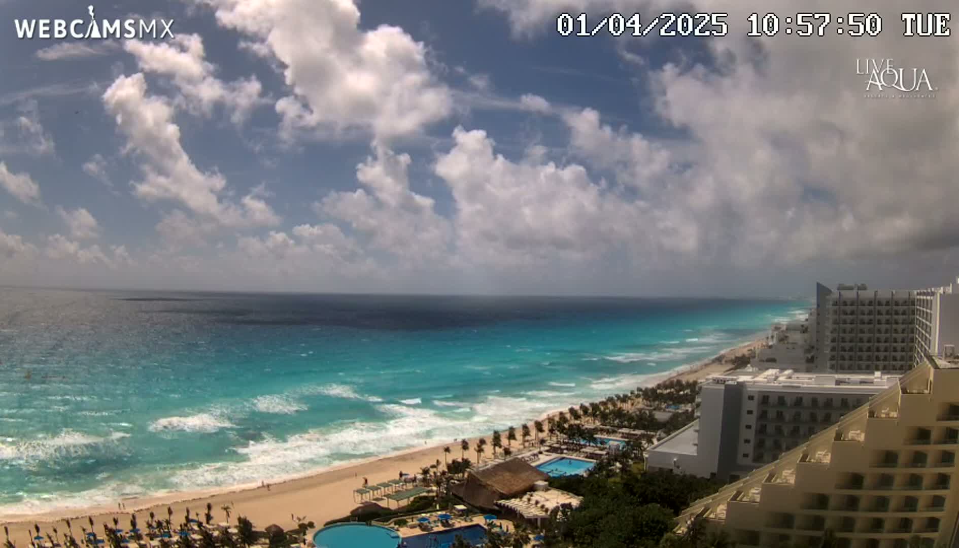 A partly cloudy day shows turquoise ocean waves breaking on a sandy beach lined with resort hotels.