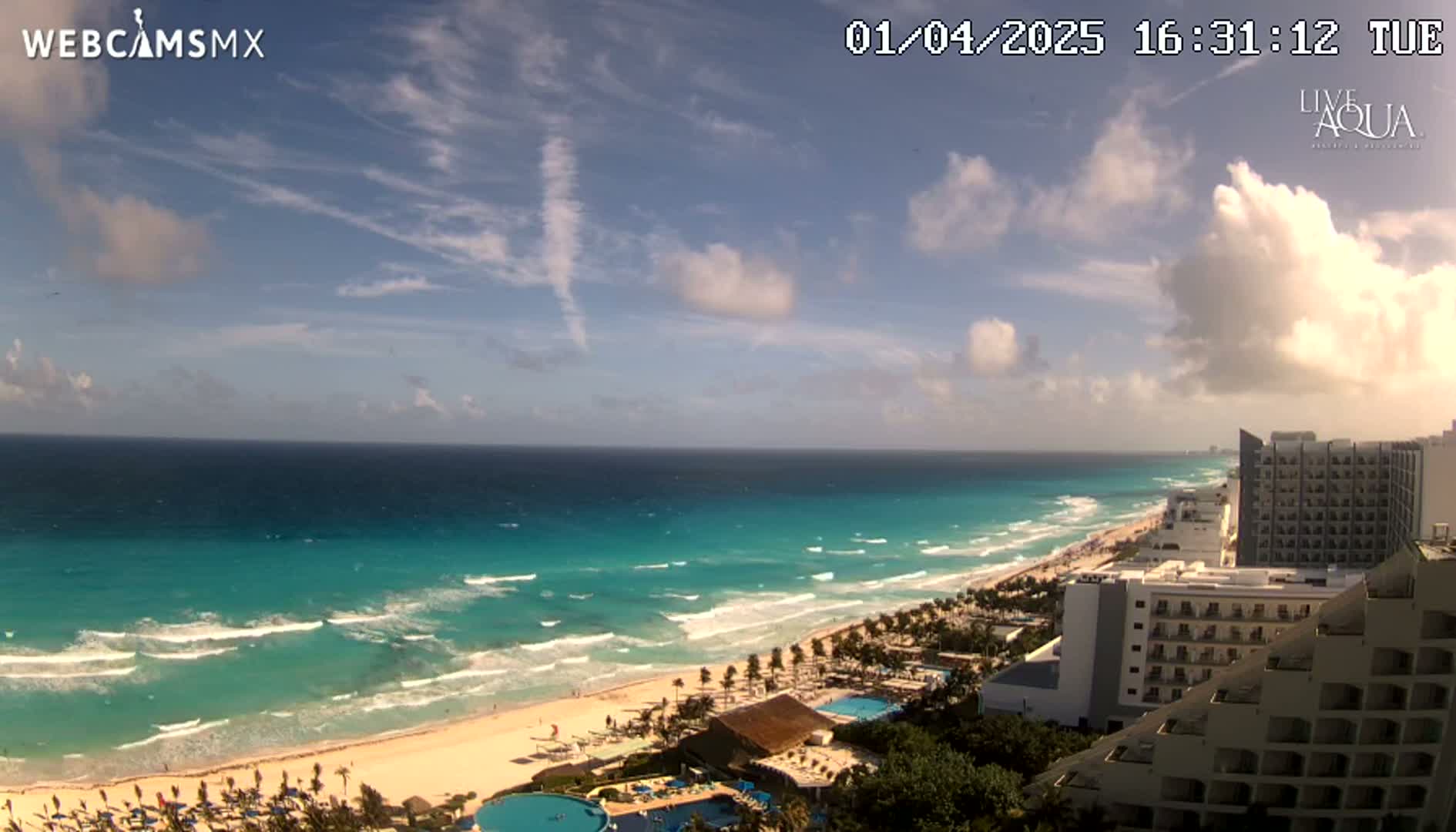 A partly cloudy sky overlooks a turquoise ocean with waves breaking on a white sandy beach lined with hotels and palm trees.
