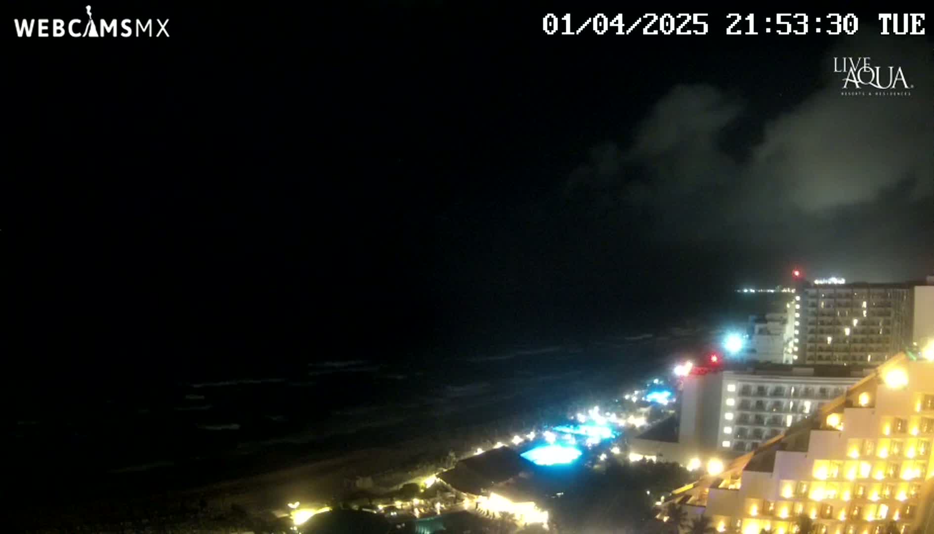 A nighttime view of a coastline with dark clouds overhead, showing a dark ocean, a sandy beach, and brightly lit hotels.