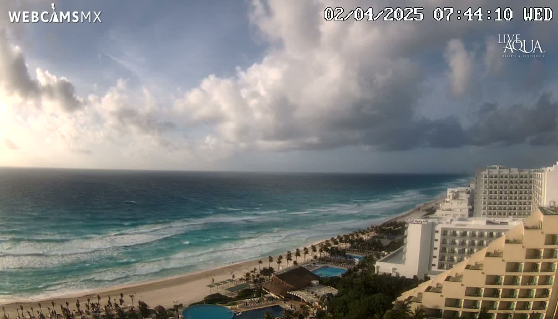 A partly cloudy sky overlooks a beach with turquoise water and waves, along with several hotels and resorts.