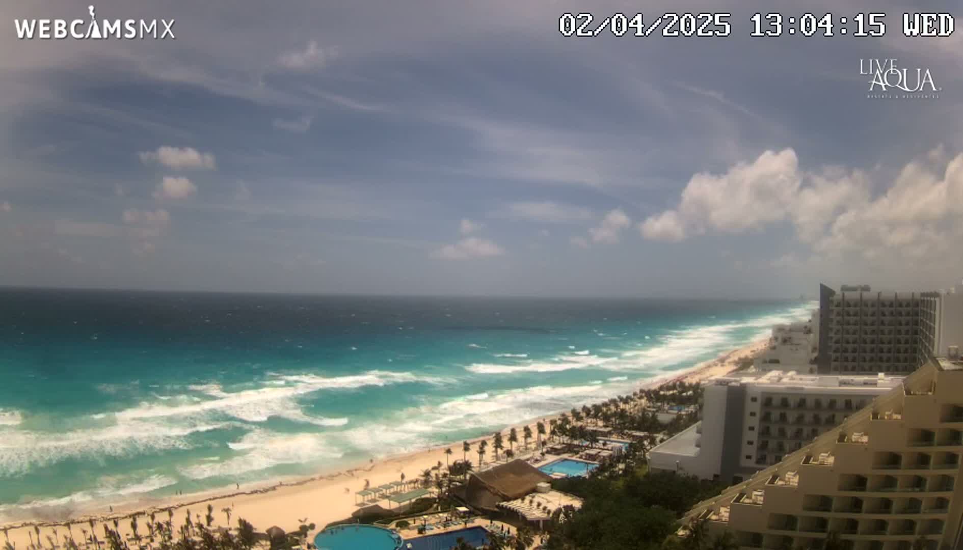 A partly cloudy day shows a beach with turquoise water and moderate waves, alongside a line of hotels.