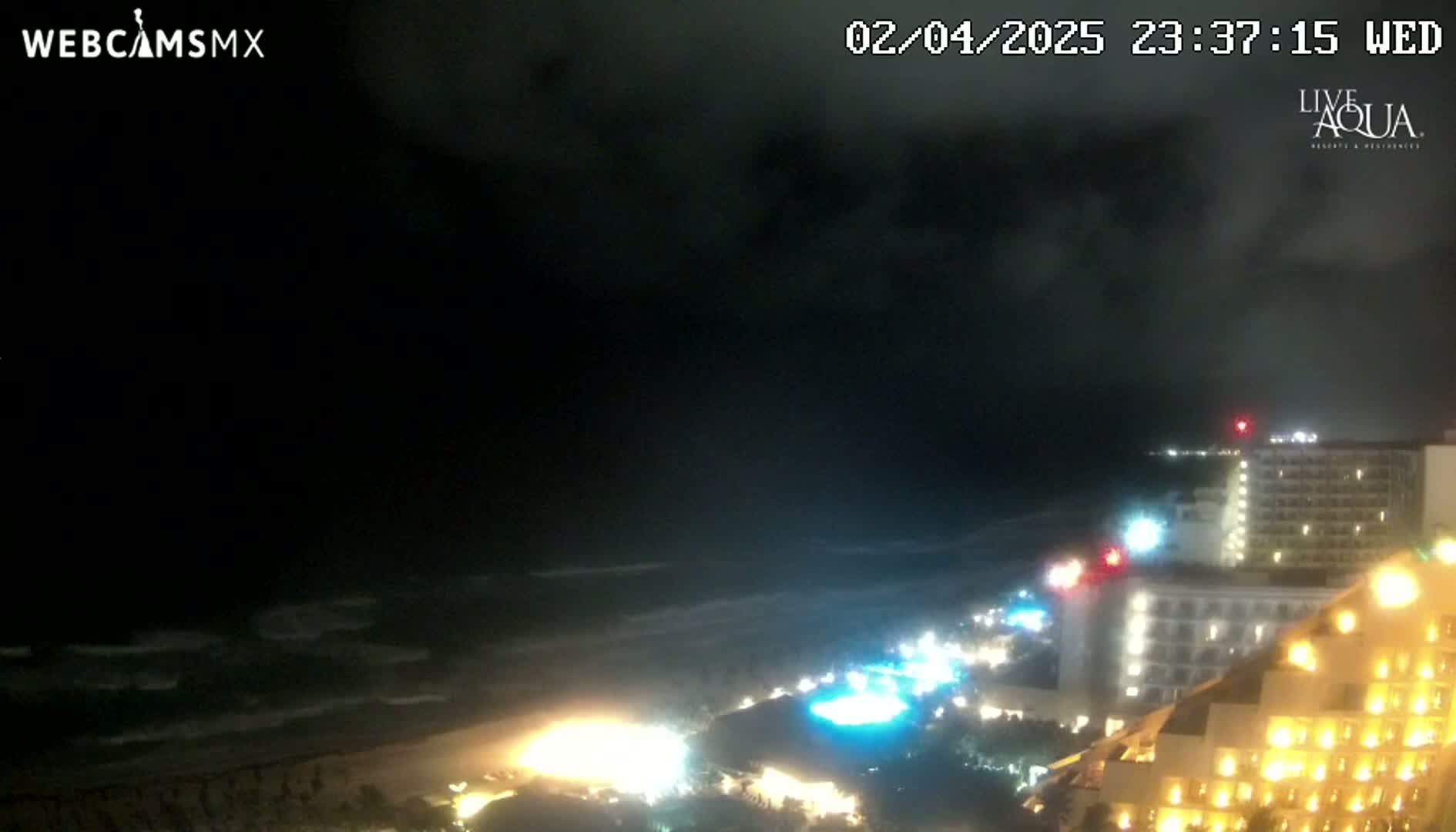 A nighttime view of a beach with dark, cloudy skies, a gently-choppy ocean, and illuminated buildings along the shore.