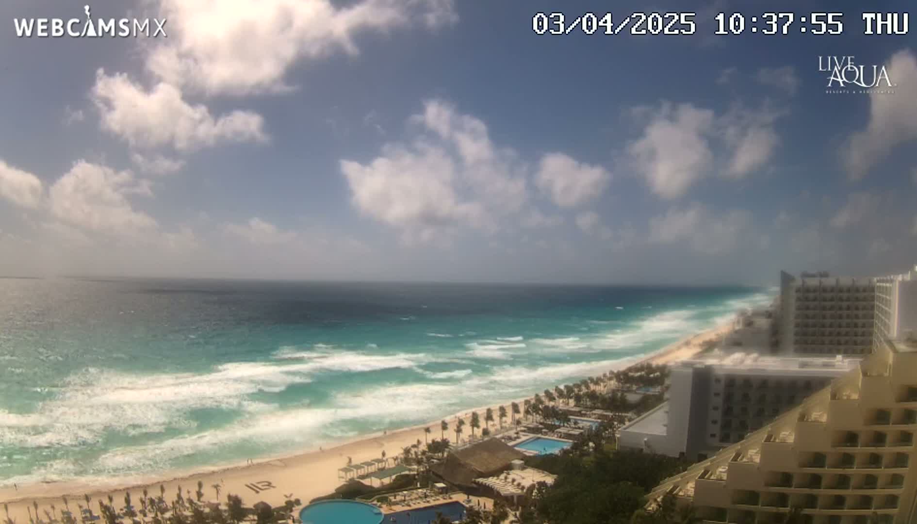 A partly cloudy day shows a beach with turquoise water and waves crashing on the shore, with resort hotels visible along the coastline.