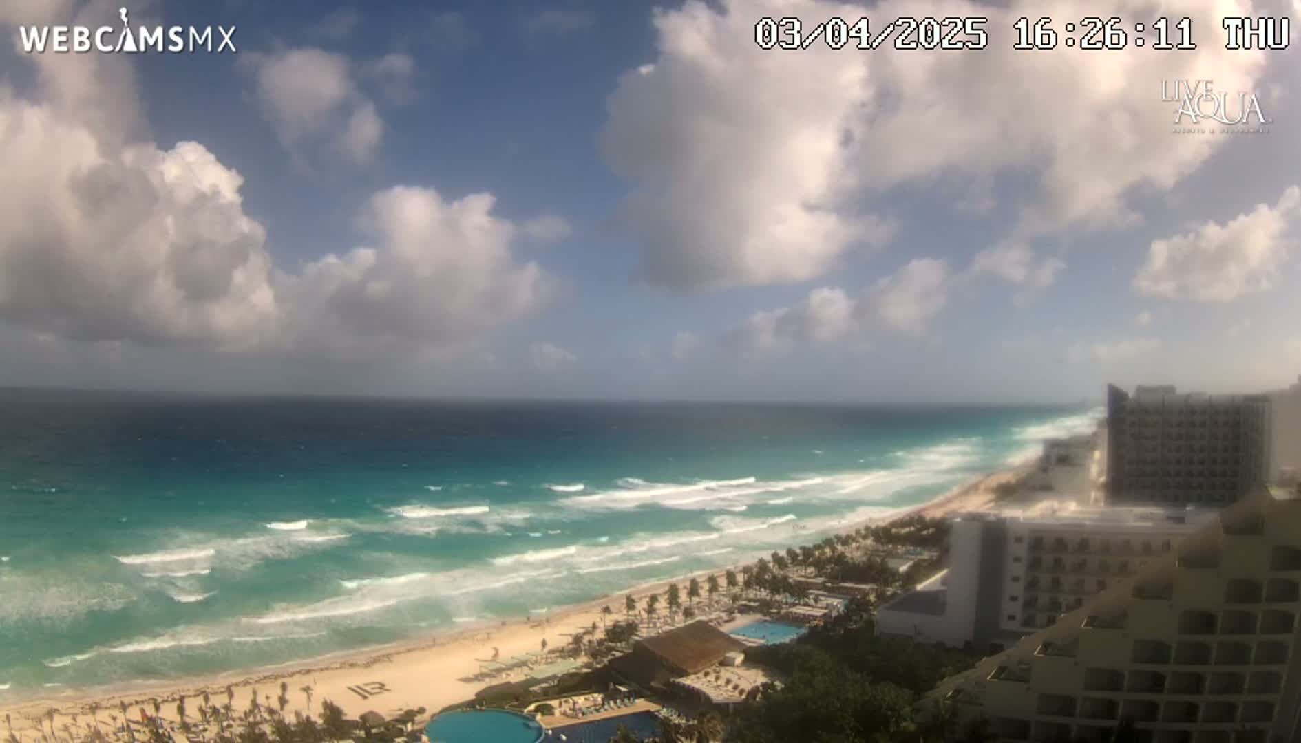 A partly cloudy day shows a turquoise ocean with waves breaking on a sandy beach, lined with hotels and resort buildings.