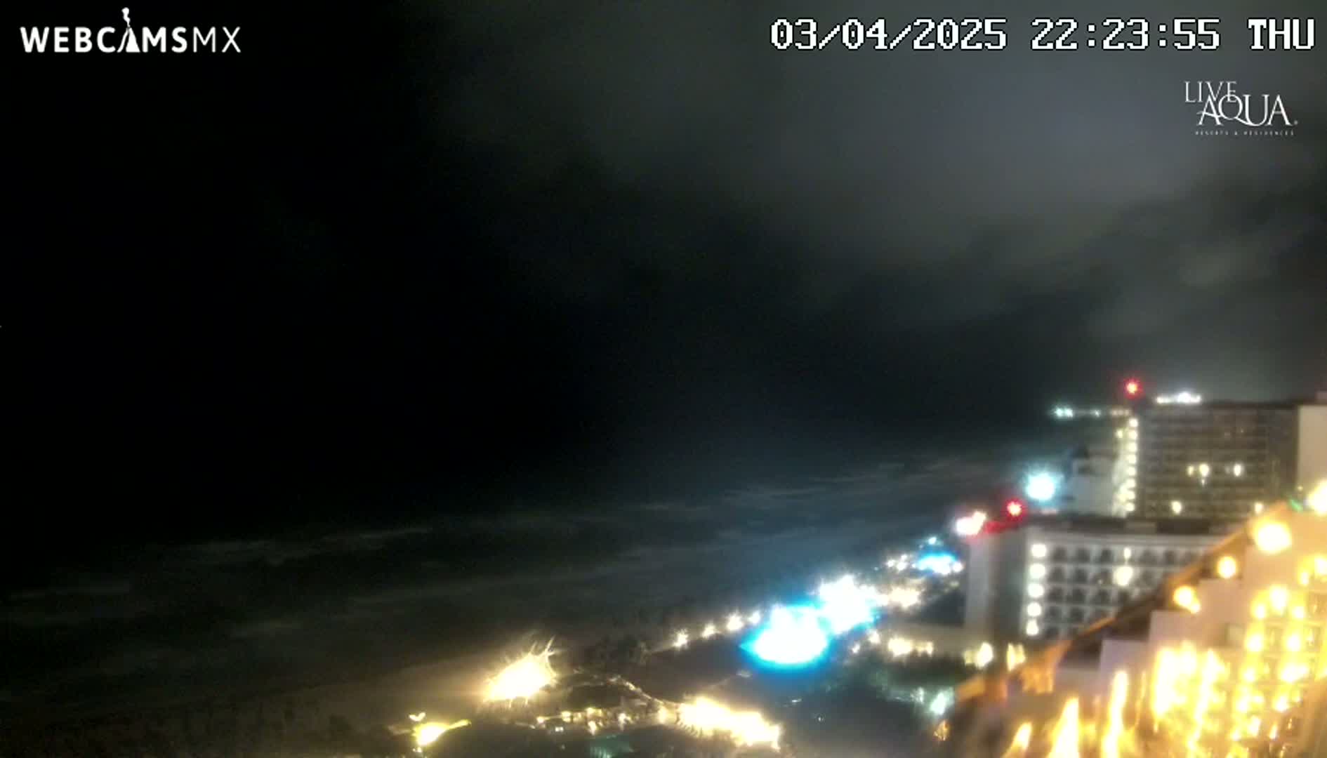 A nighttime coastal scene shows a dark, overcast sky over a wavy ocean and illuminated beachfront buildings.