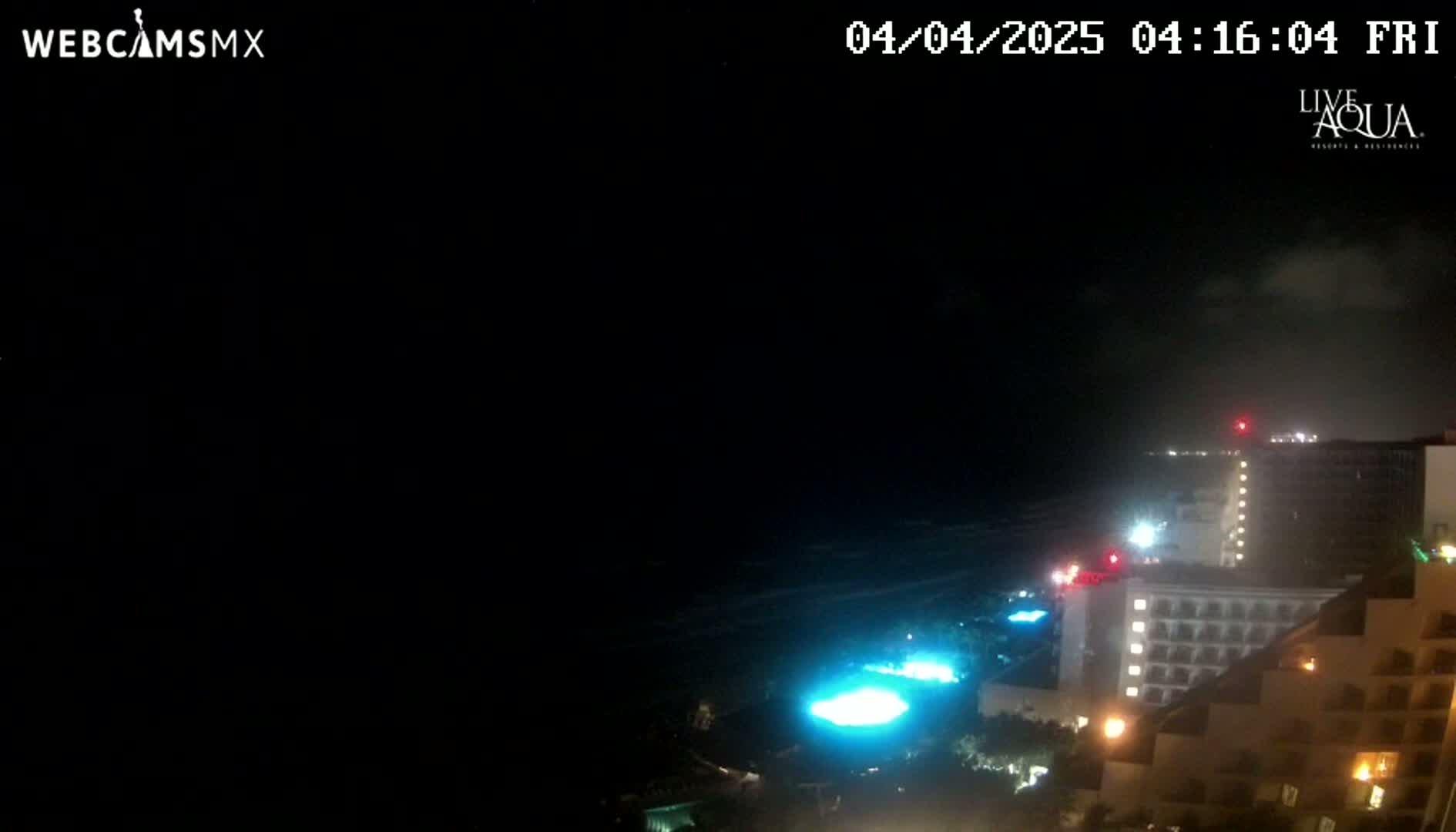 A nighttime, dark-sky view of a coastal area shows illuminated buildings and pools near a dark ocean.