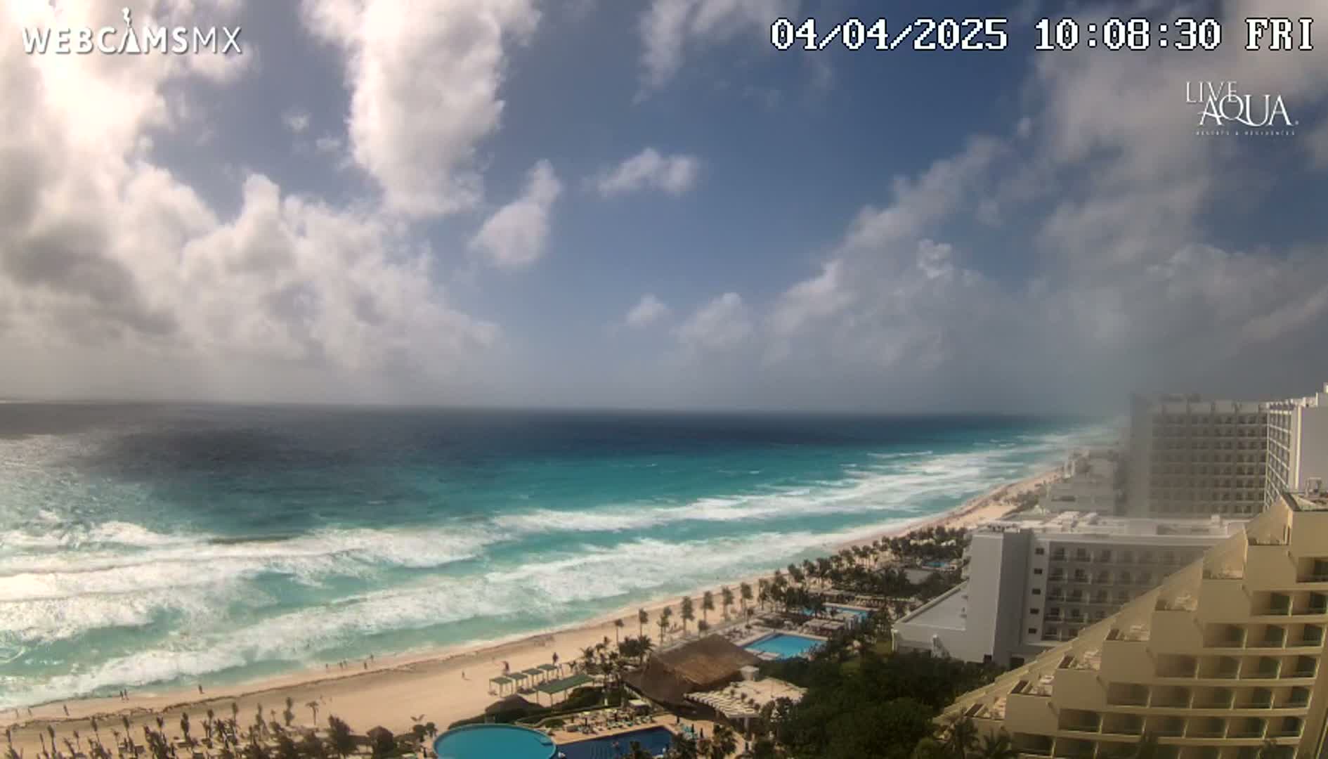 A partly cloudy day shows a beach with turquoise water and waves, along with several resort hotels.