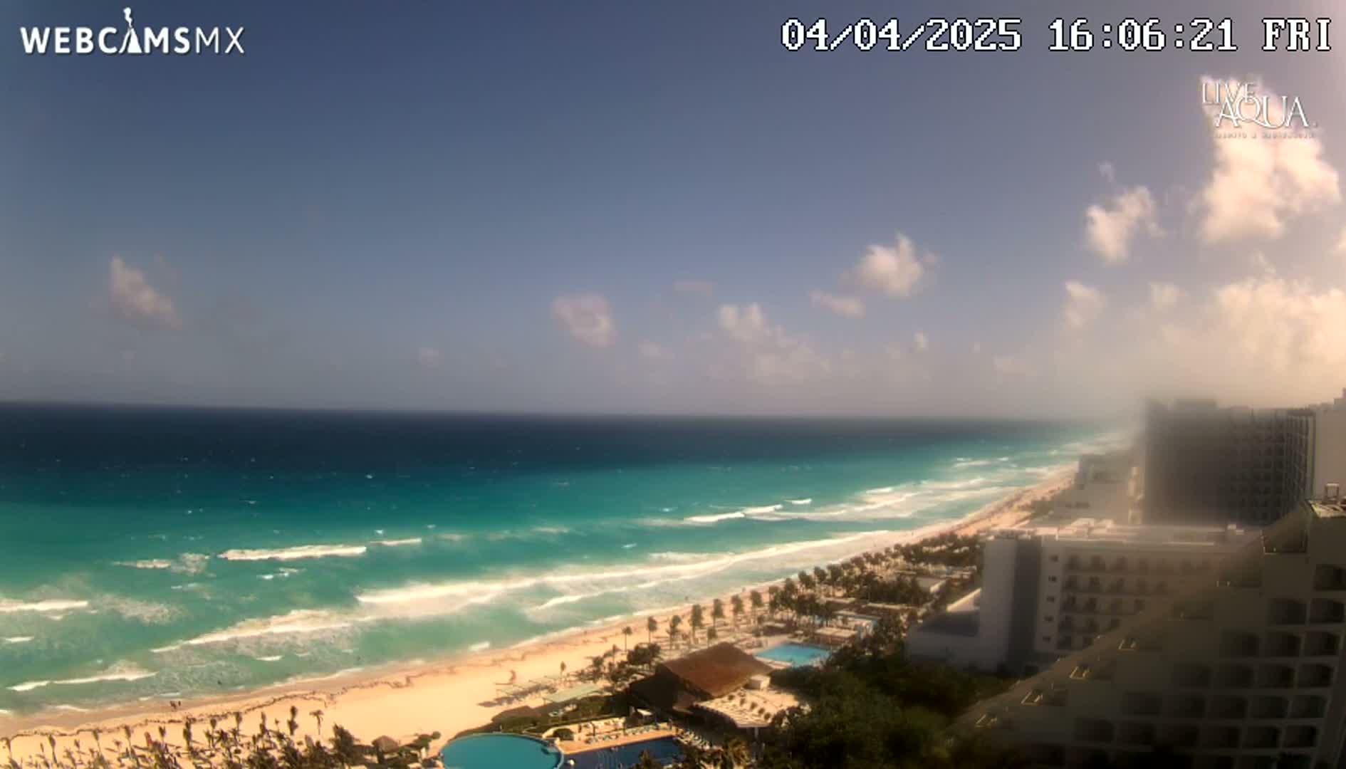 A partly cloudy day shows a long stretch of beach with turquoise water and waves, lined with hotels and resorts.