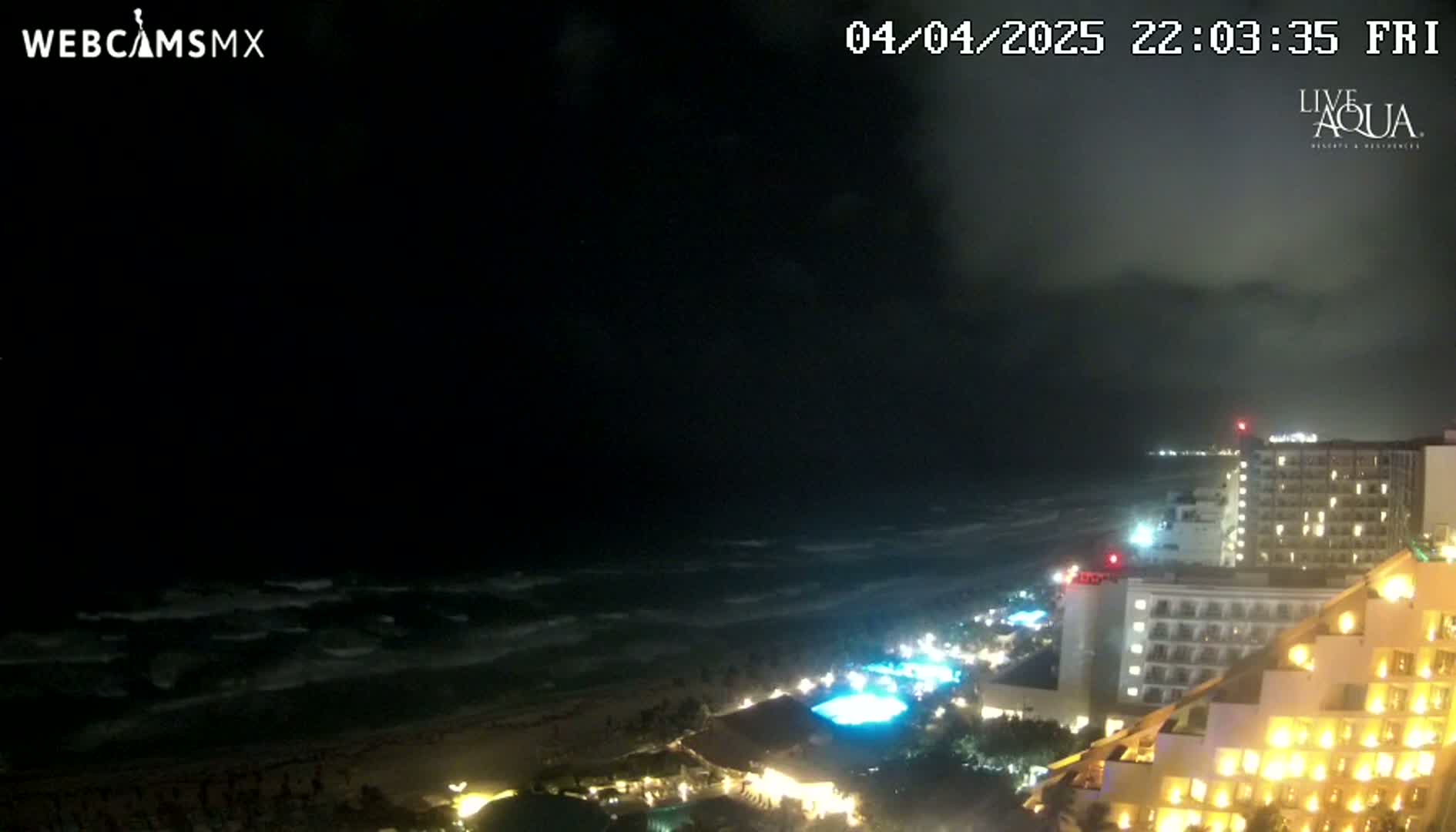 A nighttime view of a beach with moderately rough waves, illuminated hotels, and a partly cloudy sky.