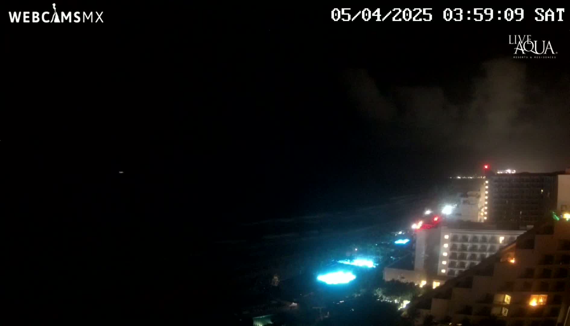 A nighttime view of a coastline shows a dark ocean, illuminated buildings, and a partly cloudy sky.