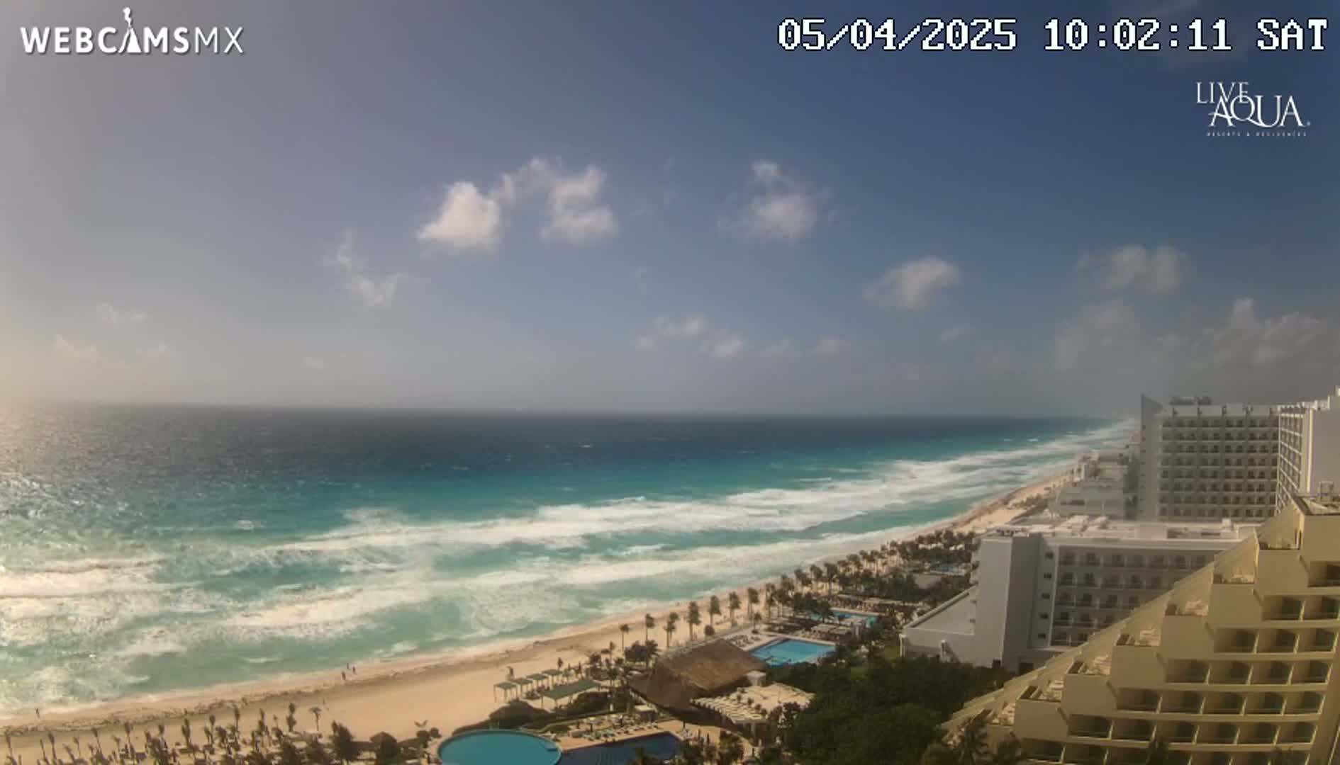 A mostly sunny day reveals a beach with moderate waves, hotels, and swimming pools overlooking the turquoise ocean.