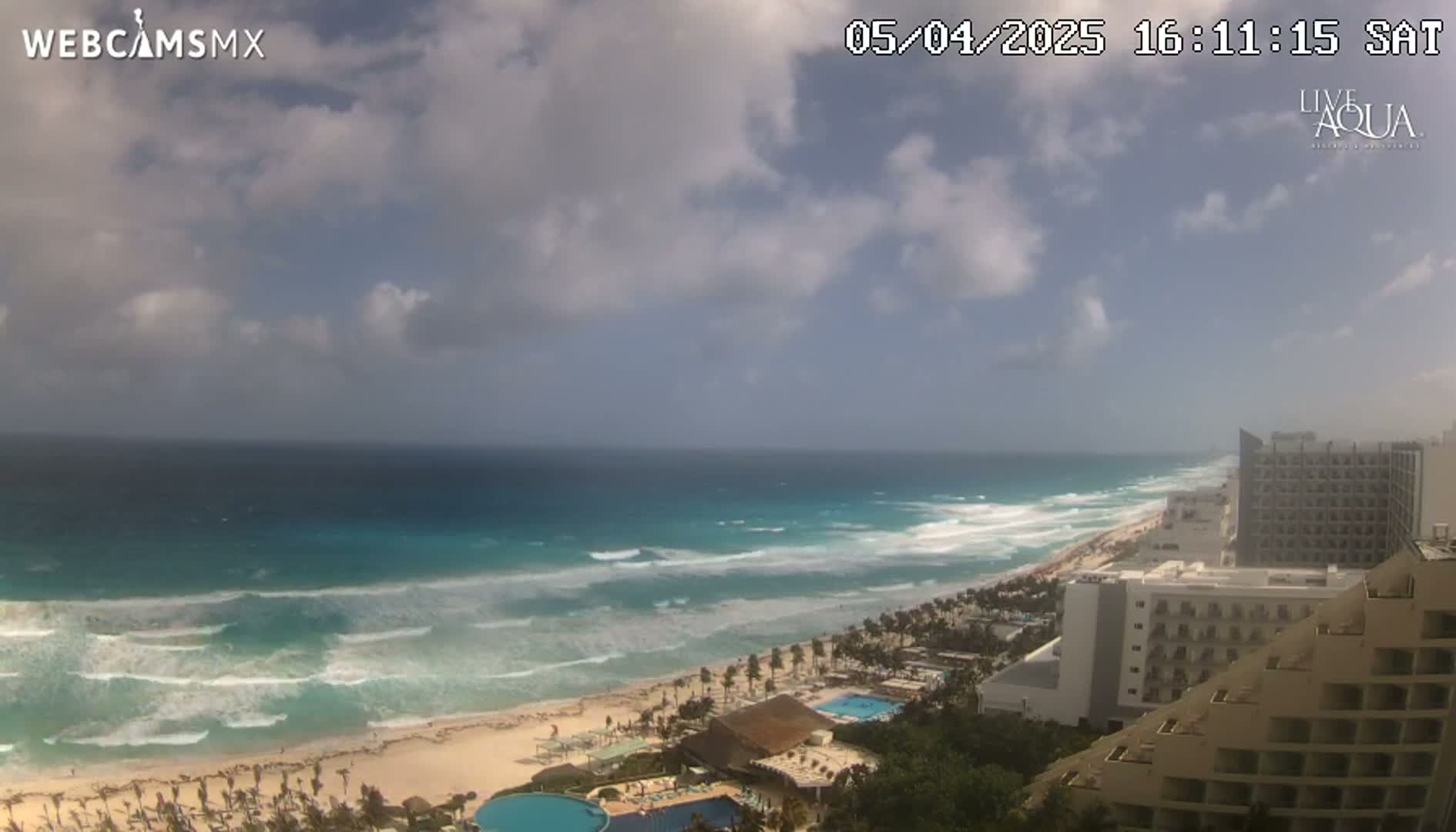 A partly cloudy day shows a wide expanse of ocean with moderate waves breaking on a sandy beach lined with resort hotels.