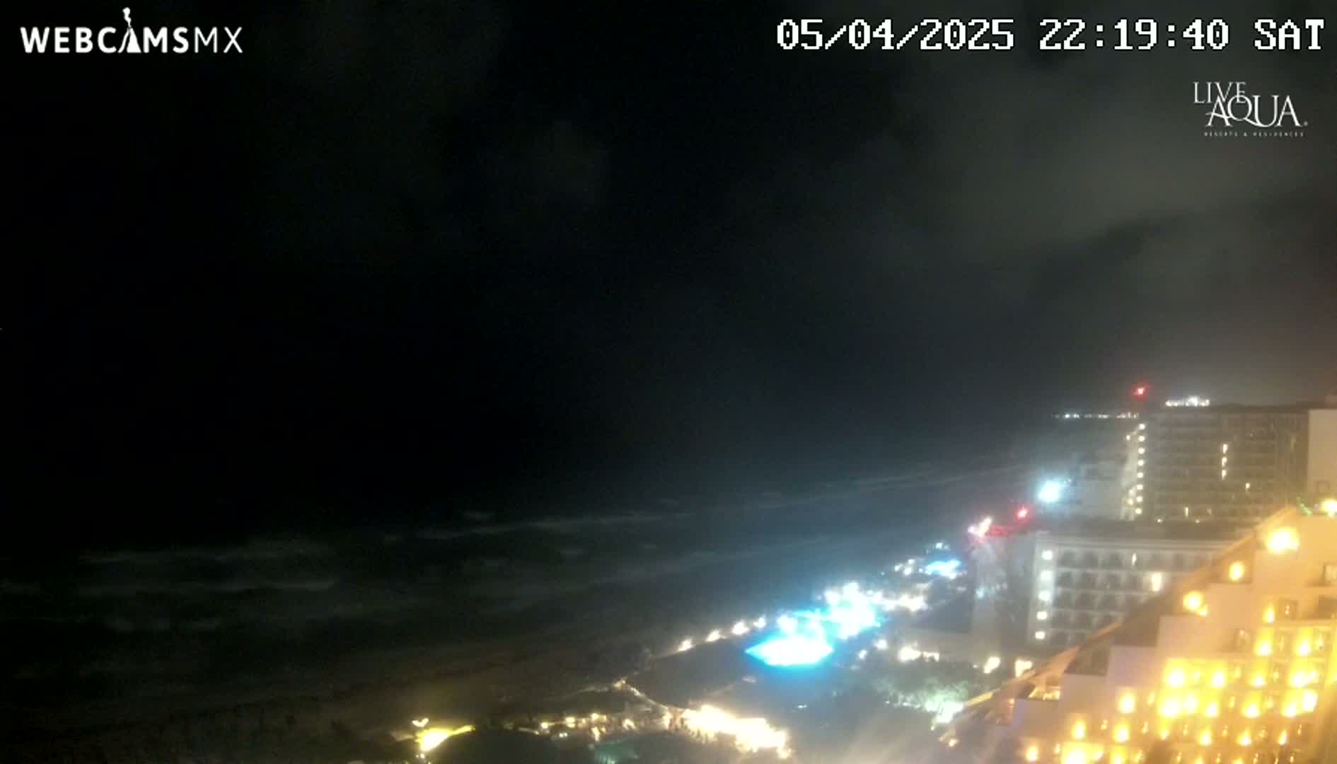 A nighttime view of a coastline shows dark, wavy ocean water and brightly lit hotels along the shore under a partly cloudy sky.