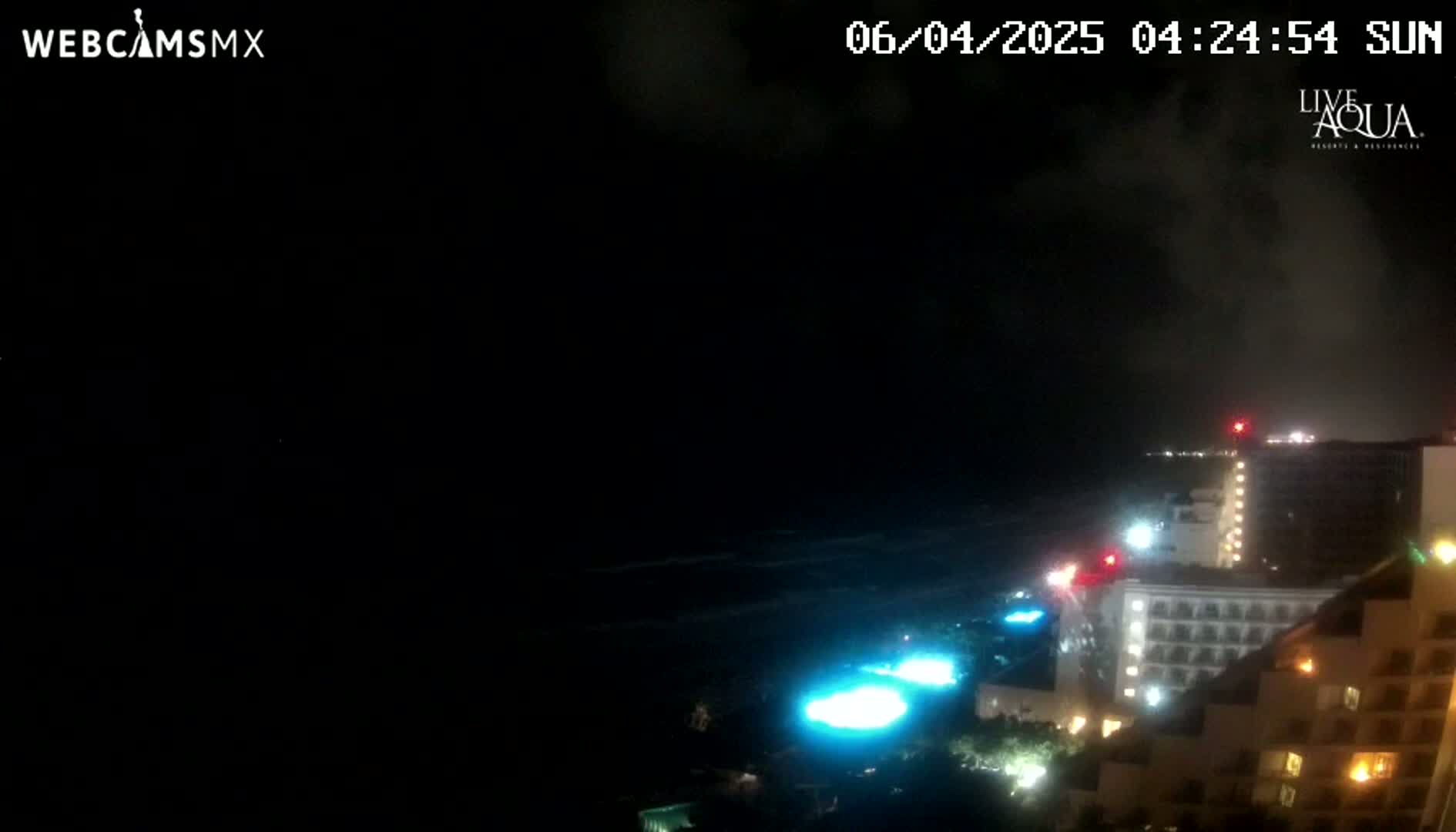 A nighttime coastal scene shows a dark ocean with waves under a cloudy sky, illuminated by the lights of several large buildings along the shore.
