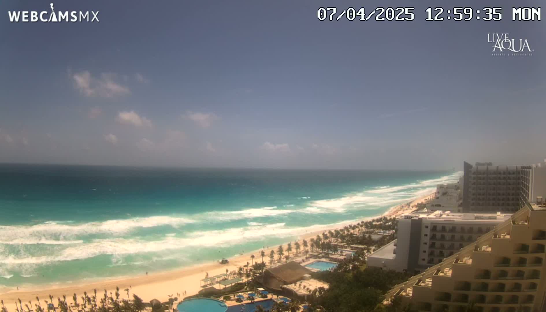A partly cloudy day shows turquoise water with white waves crashing on a sandy beach lined with resorts.