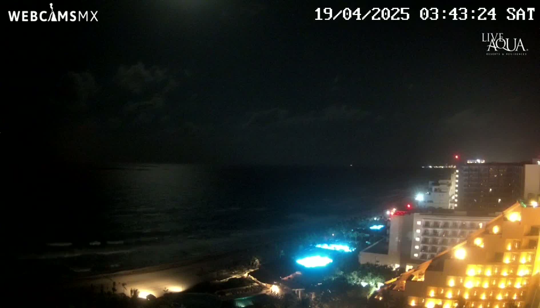 A nighttime view of a beach with a dark ocean under a partly cloudy sky, overlooking illuminated hotels and resort pools.