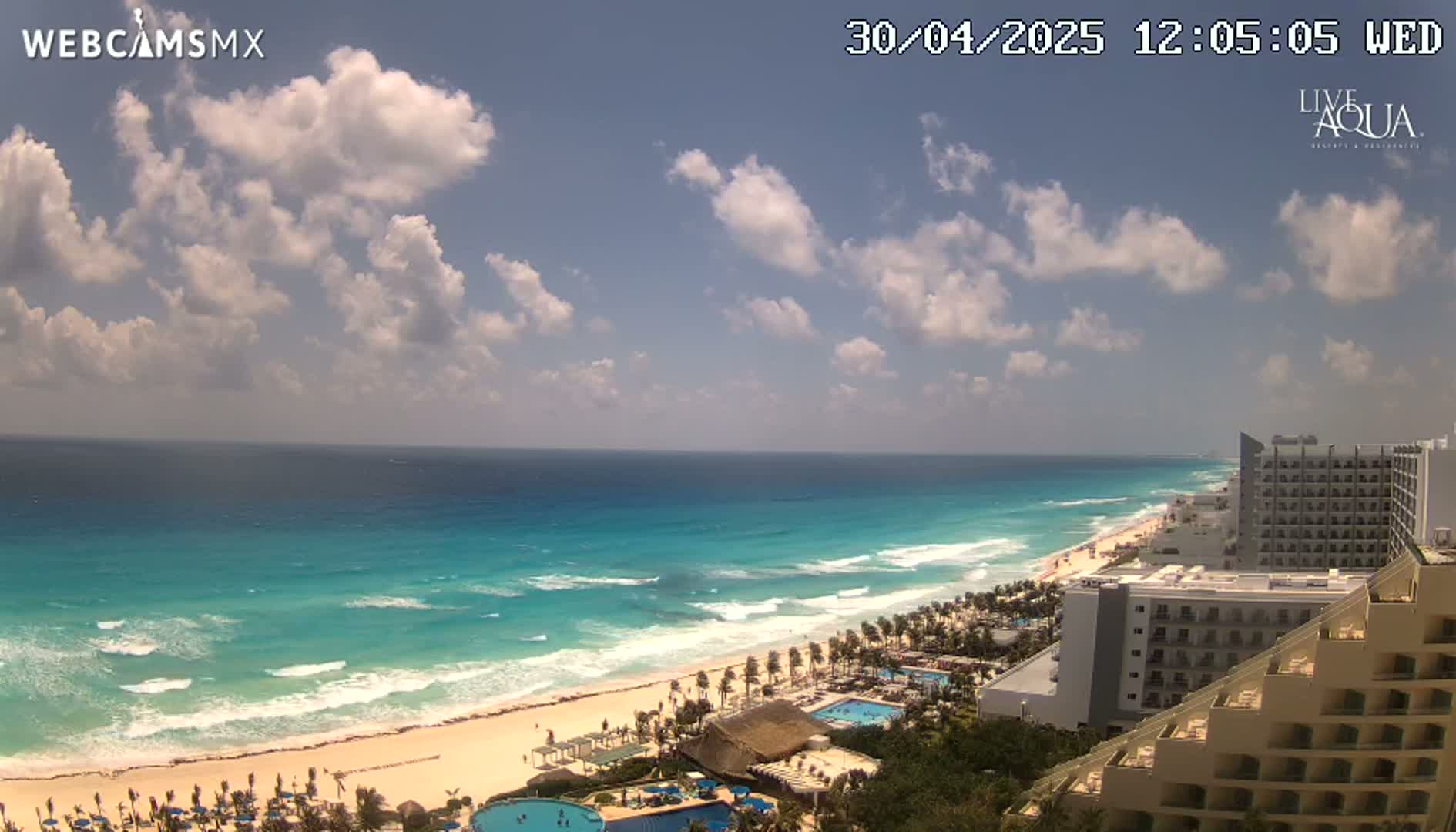 A partly cloudy day shows a long stretch of beach with turquoise water, waves, and several hotels along the shoreline.