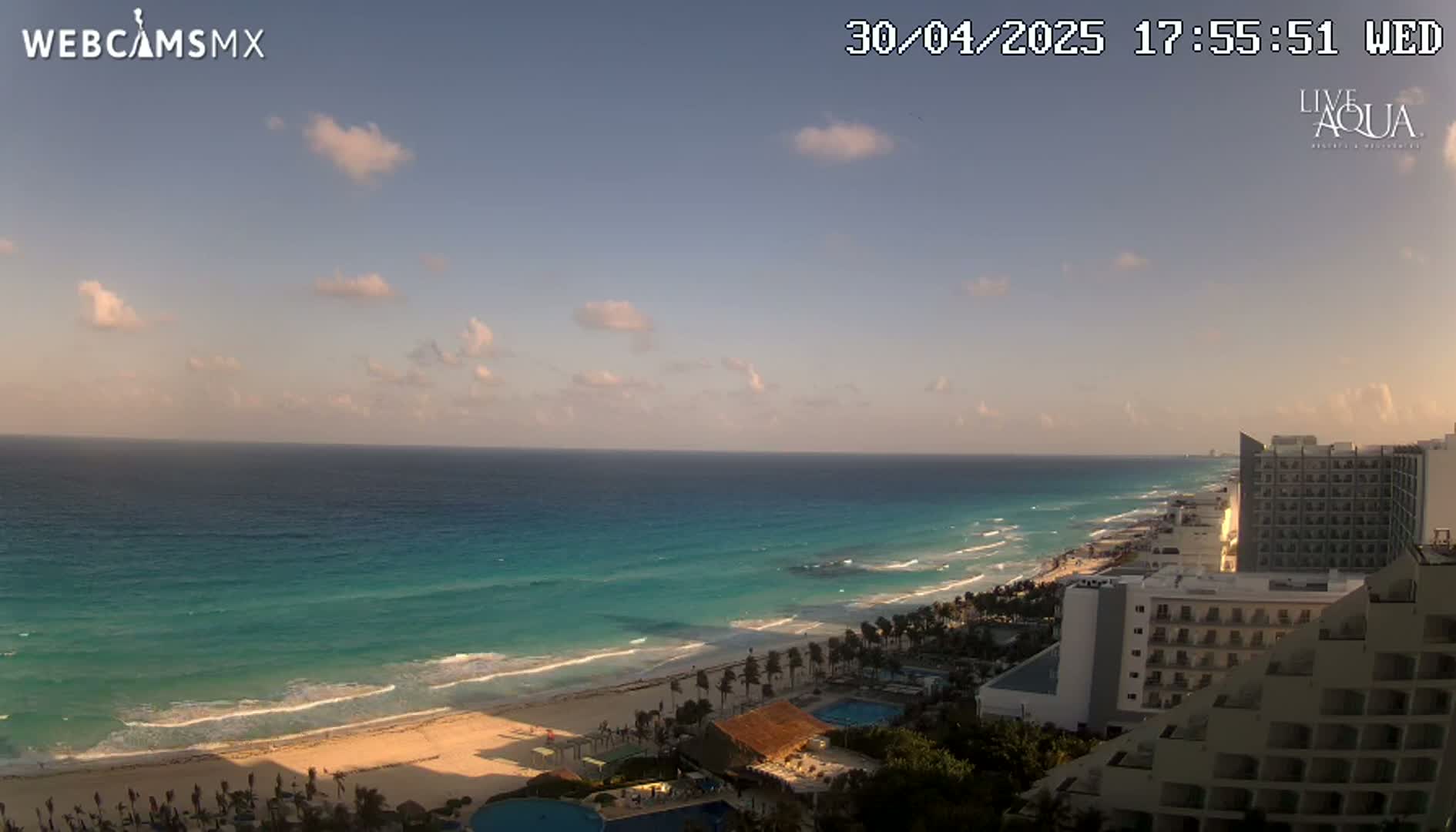 A partly cloudy sunset overlooks a turquoise ocean, sandy beach, and beachfront hotels.