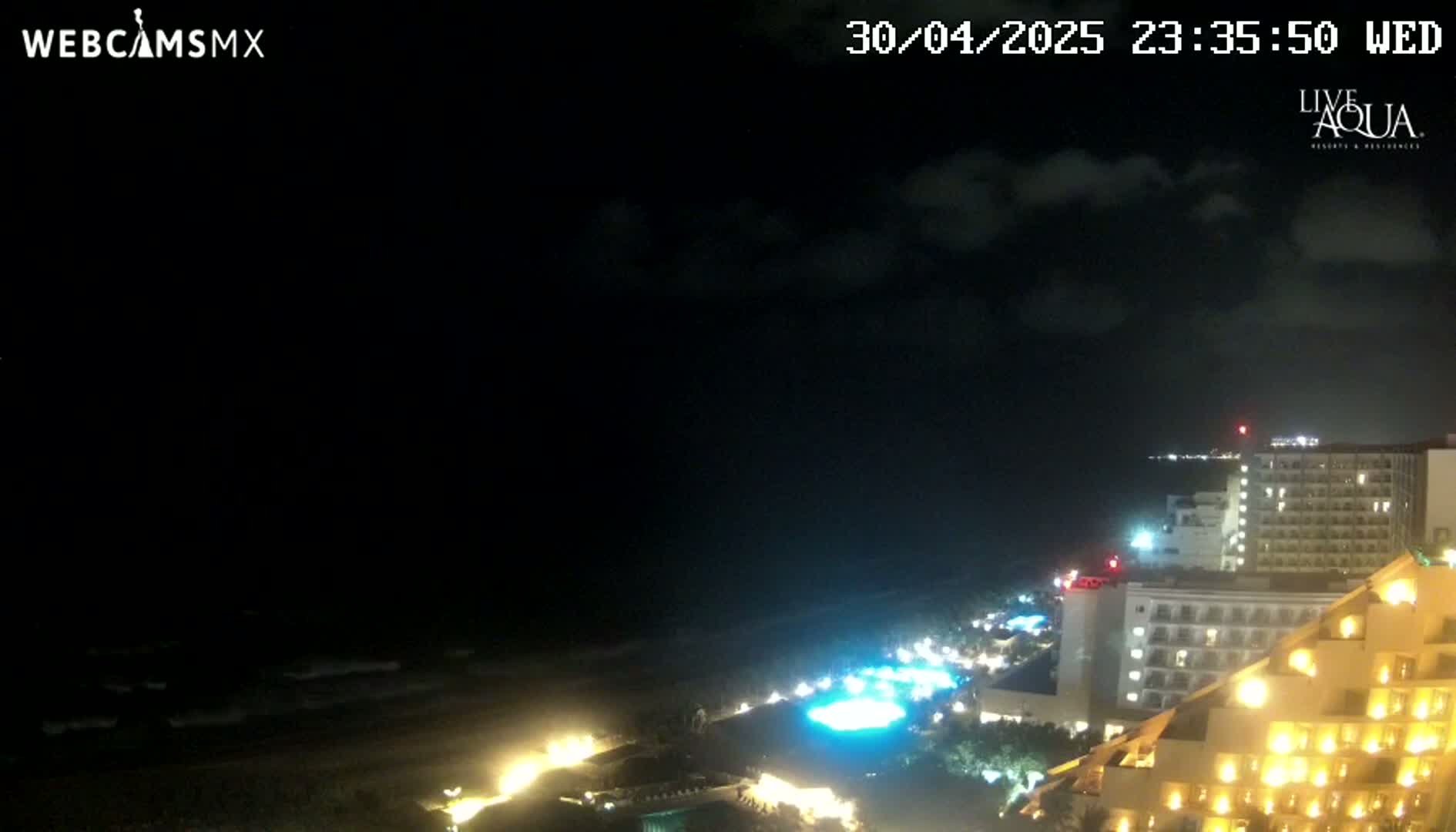 A nighttime coastal view shows a dark ocean with waves, a brightly lit beachfront resort, and a partly cloudy sky.