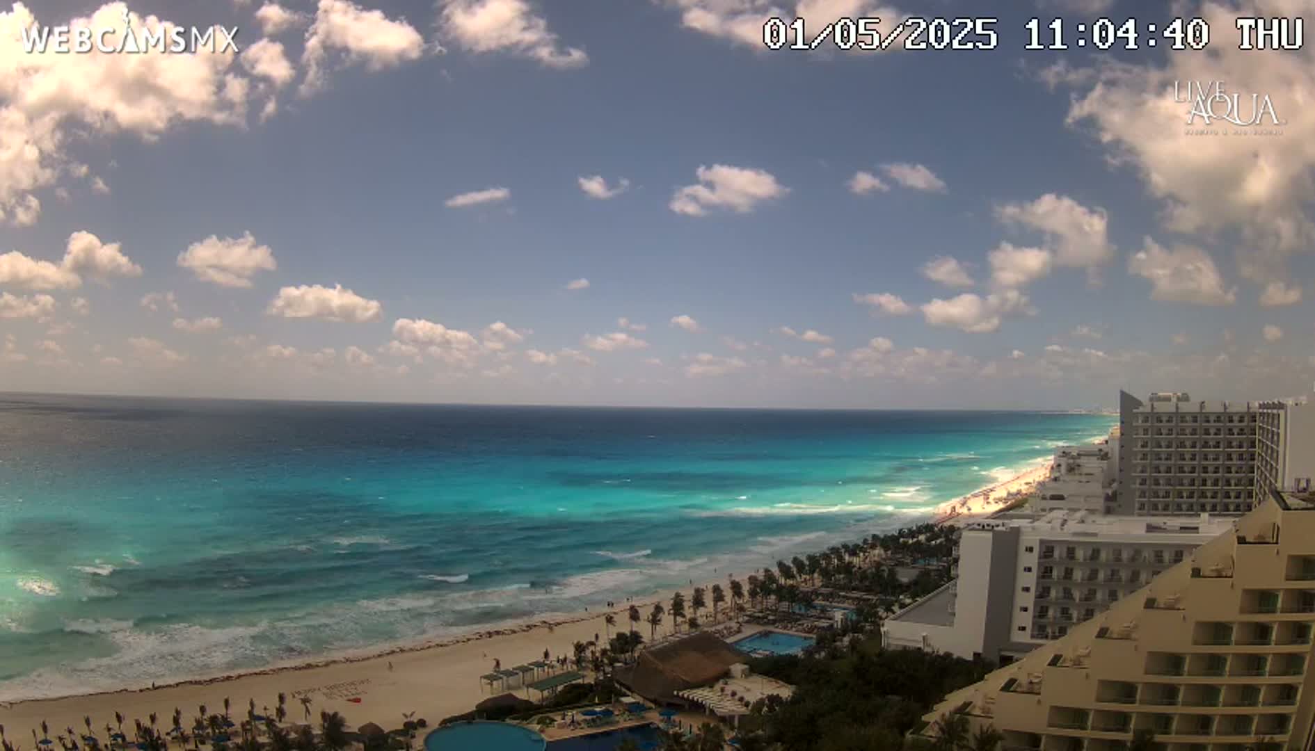 A partly cloudy day shows a beach with turquoise water, waves lapping the shore, and hotels lining the coast.
