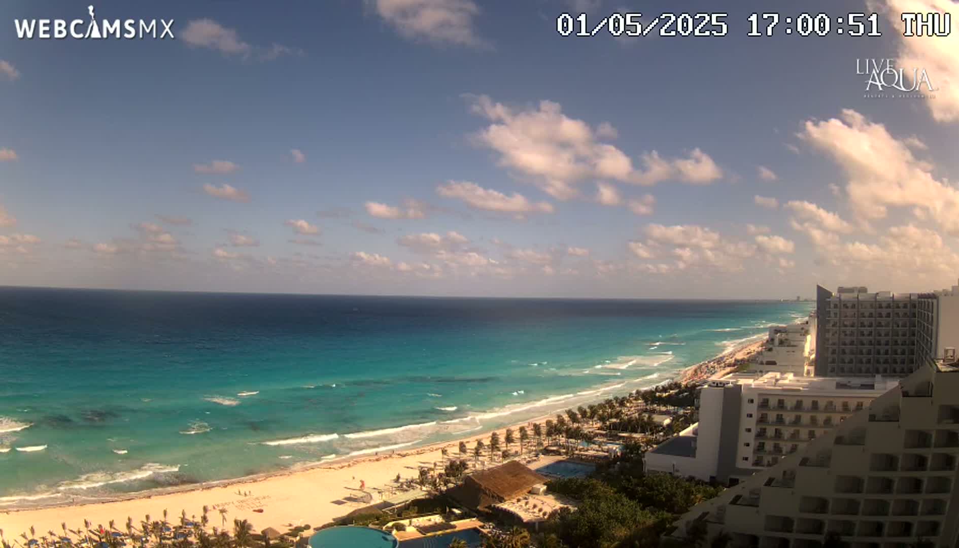 A partly cloudy sky overlooks a long, sandy beach with turquoise water lapping the shore, backed by hotels and resort buildings.