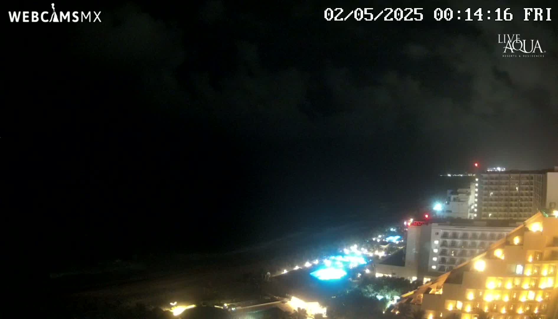 A nighttime coastal scene shows a dark ocean under a cloudy sky, with illuminated hotels and resort lights along the shore.