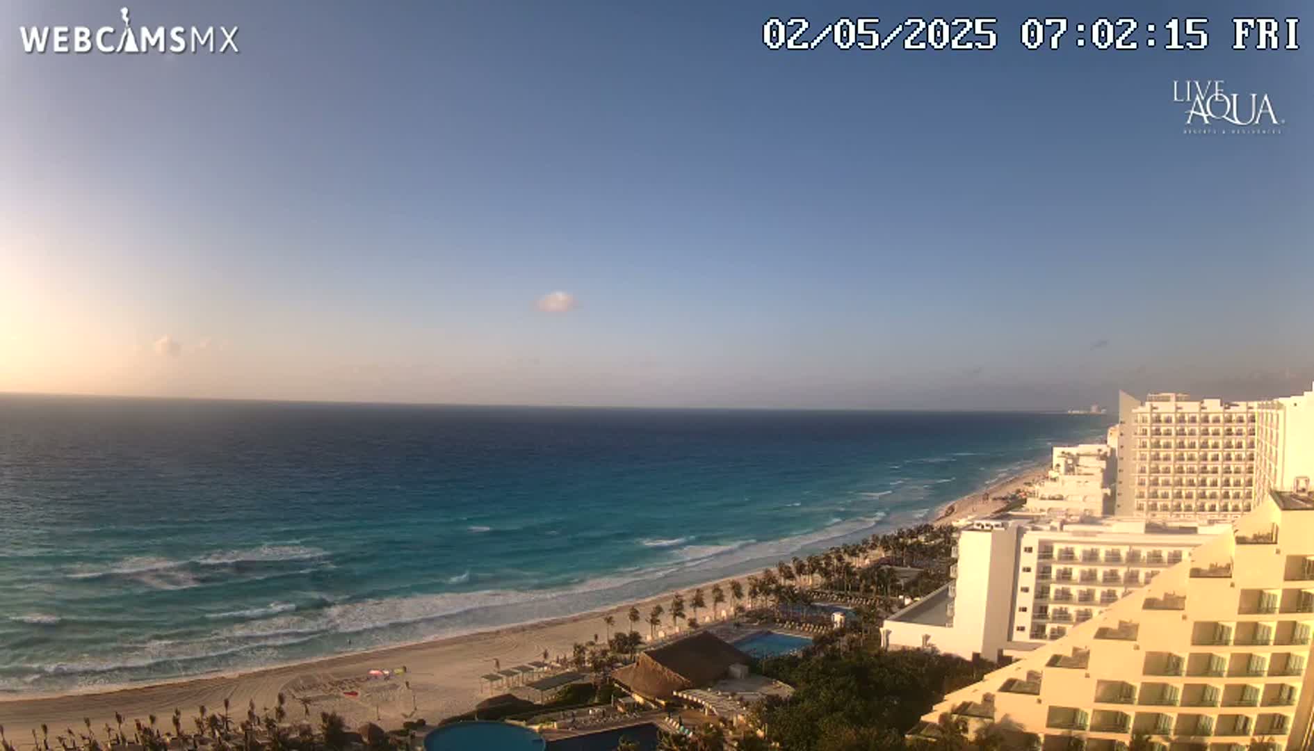 A clear sunrise illuminates a sandy beach, turquoise ocean with small waves, and numerous hotels along the coast.