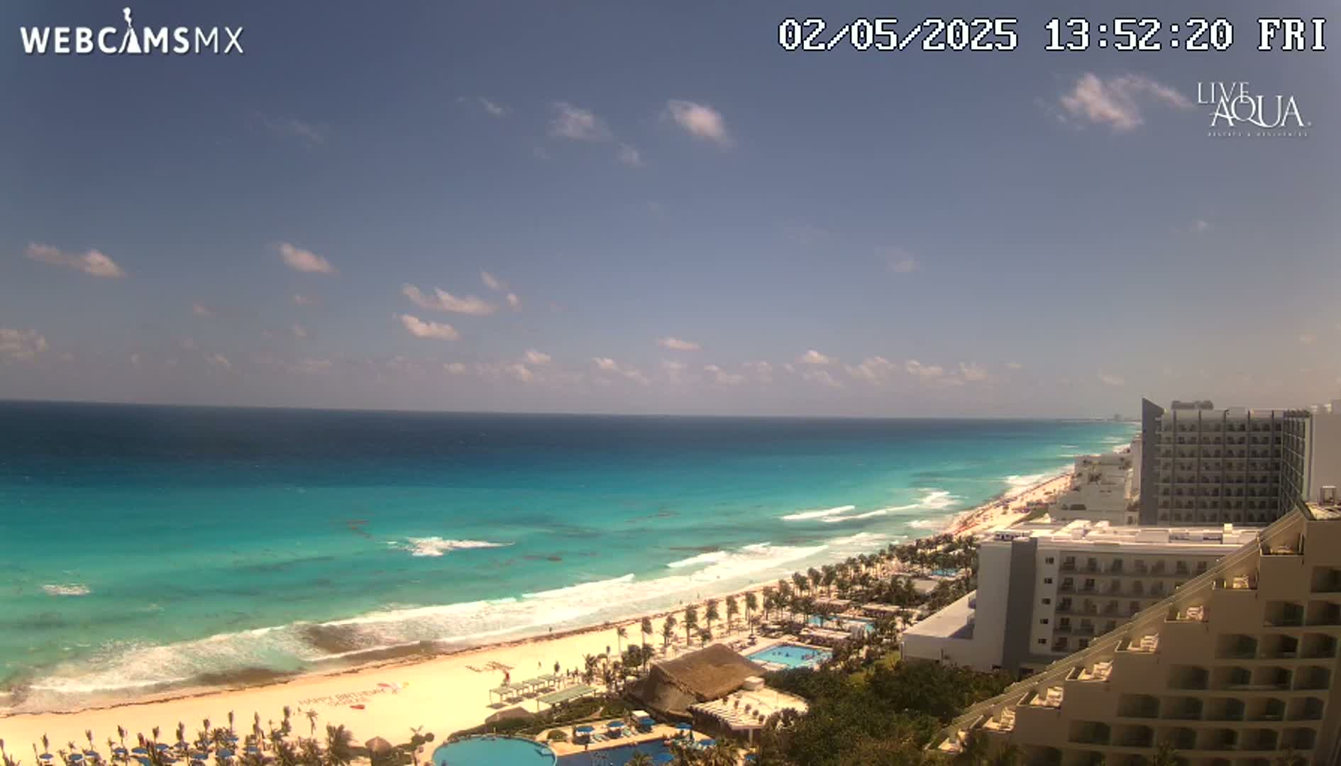 A mostly sunny day reveals a turquoise ocean meeting a white sandy beach lined with hotels and palm trees.
